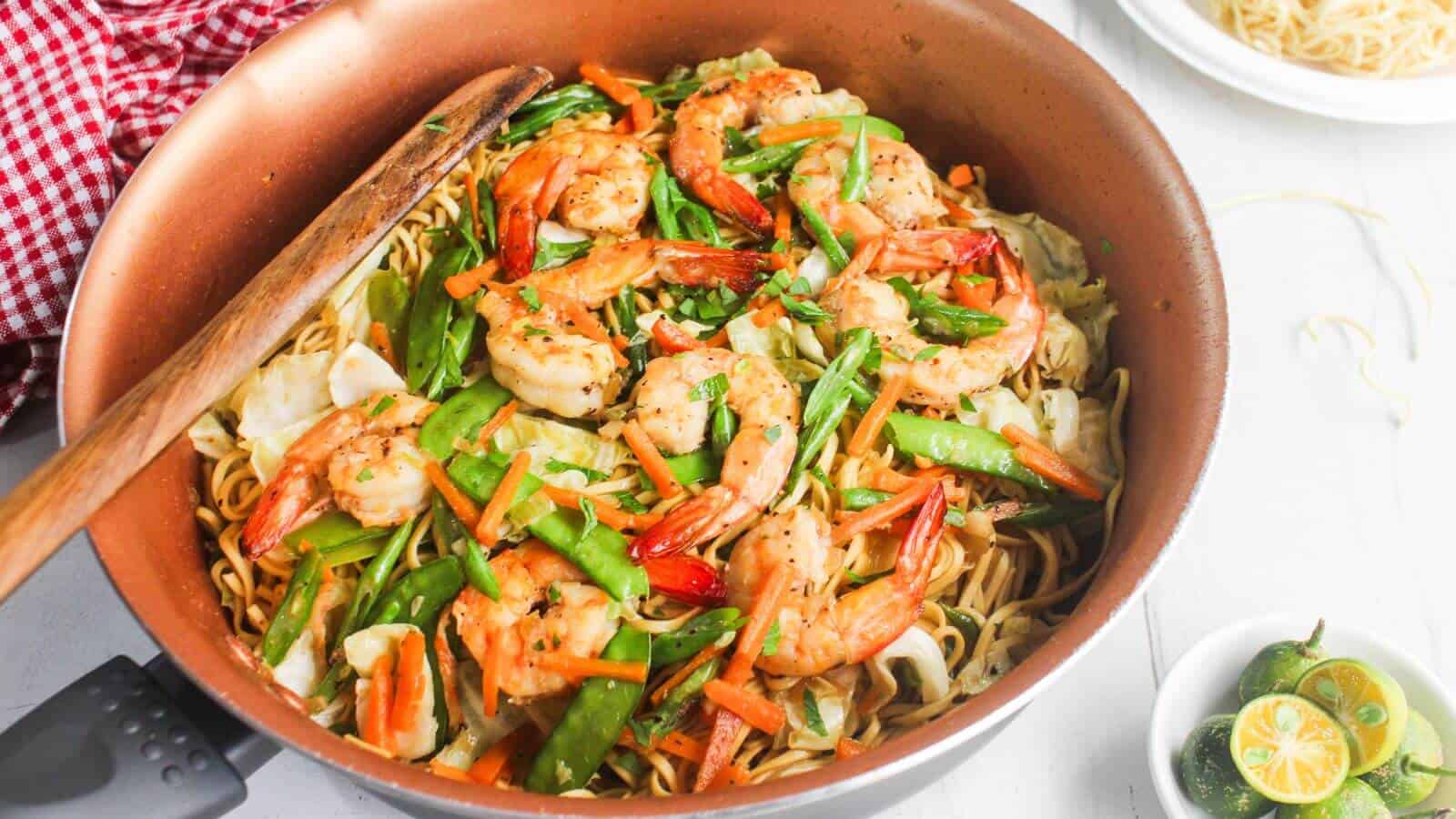 A pan of stir-fried noodles with shrimp, snap peas, carrots, and cabbage, with a wooden spatula resting on the side.