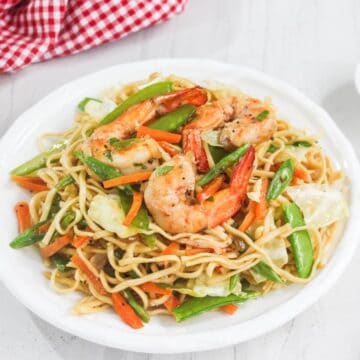 A plate of stir-fried noodles with shrimp, carrots, cabbage, and snap peas on a white table.
