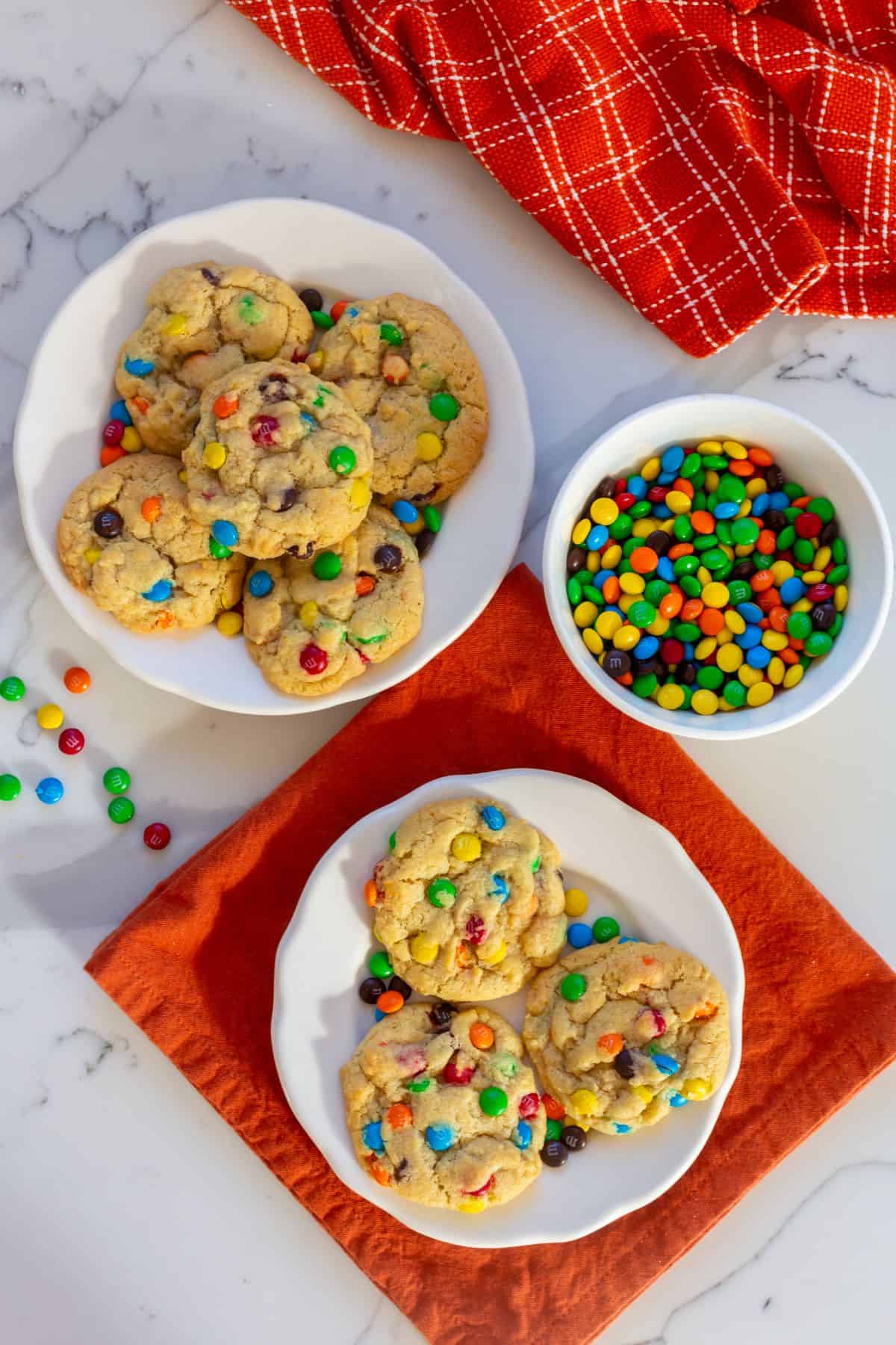 Two plates of cookies with colorful candy-coated chocolates, a bowl of the same candies, and a red cloth napkin on a white marble surface.