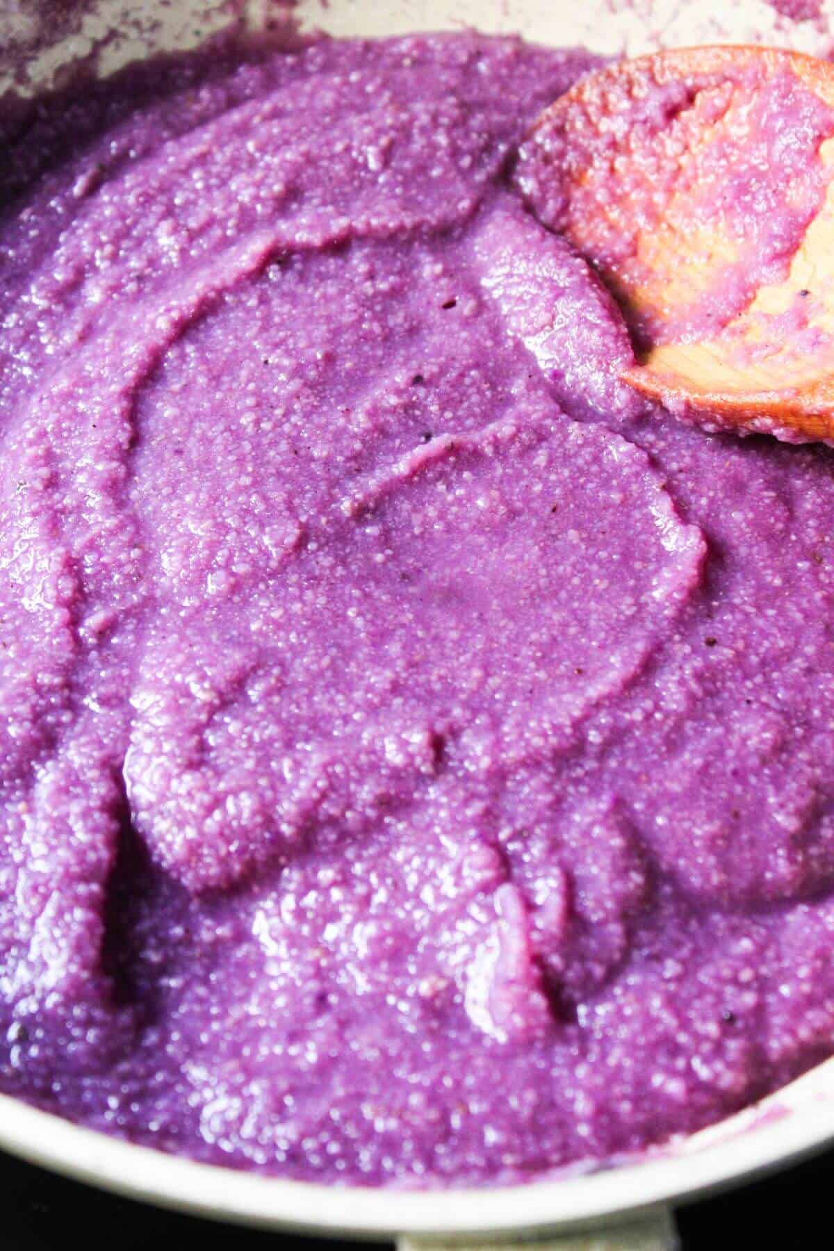 A close-up of thick, purple yam paste being stirred with a wooden spoon in a pan.