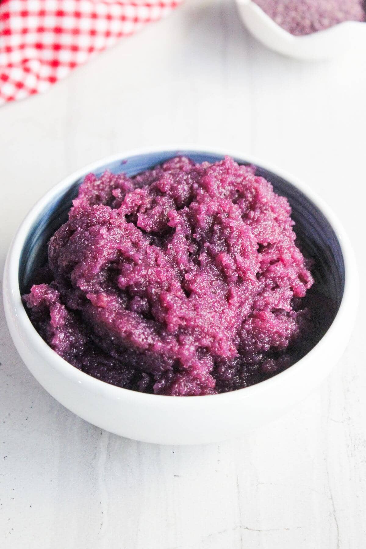 A white bowl filled with purple yam porridge, placed on a white surface with a red and white checkered cloth in the background.