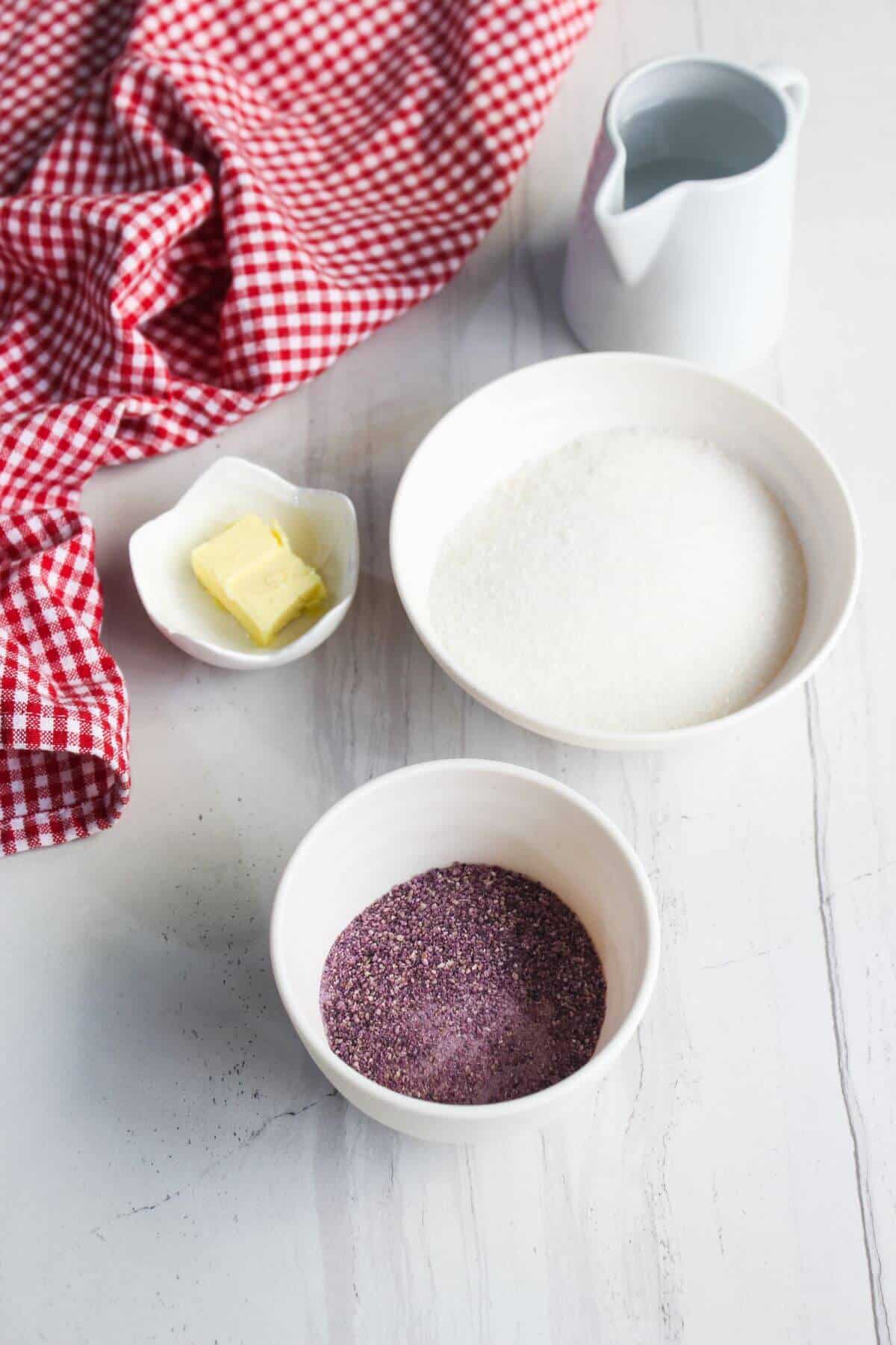 A bowl of sugar, a bowl of purple powder, a small dish of butter, and a small pitcher of water on a white surface with a red and white checkered cloth.