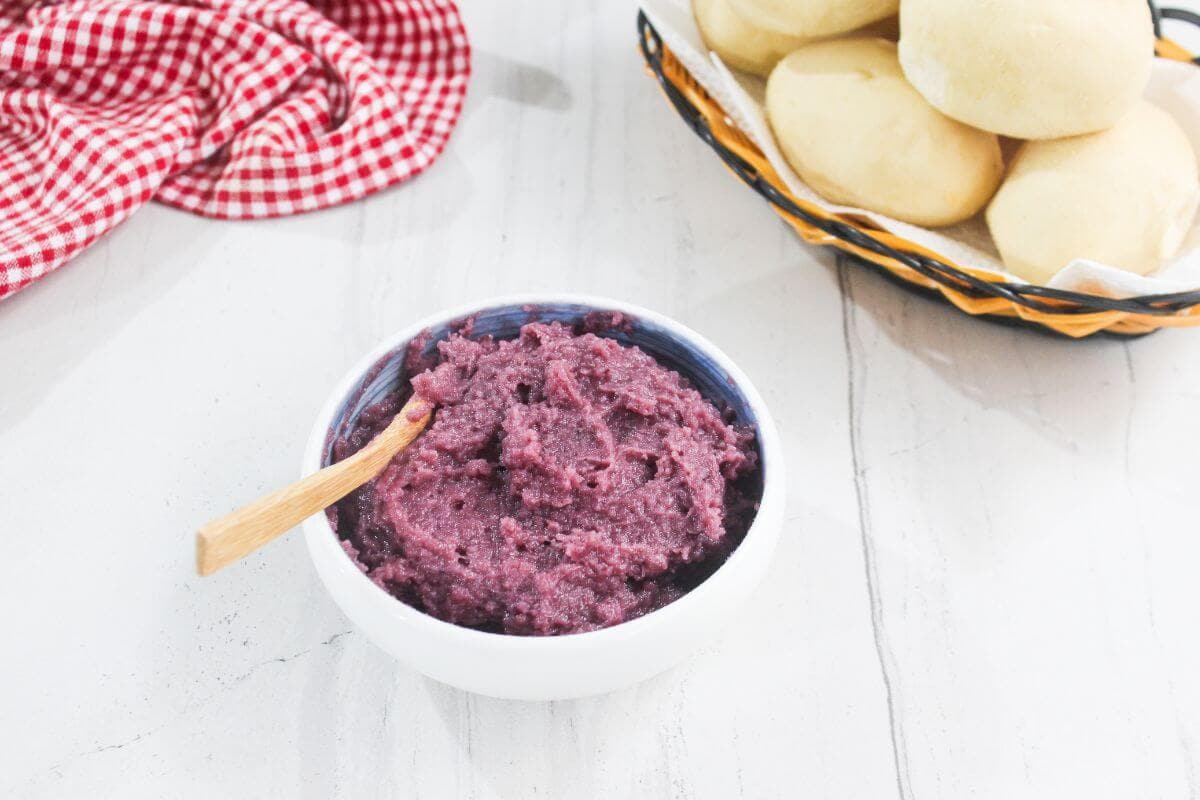A bowl of purple yam spread with a wooden spoon next to a basket of round bread rolls on a white countertop.