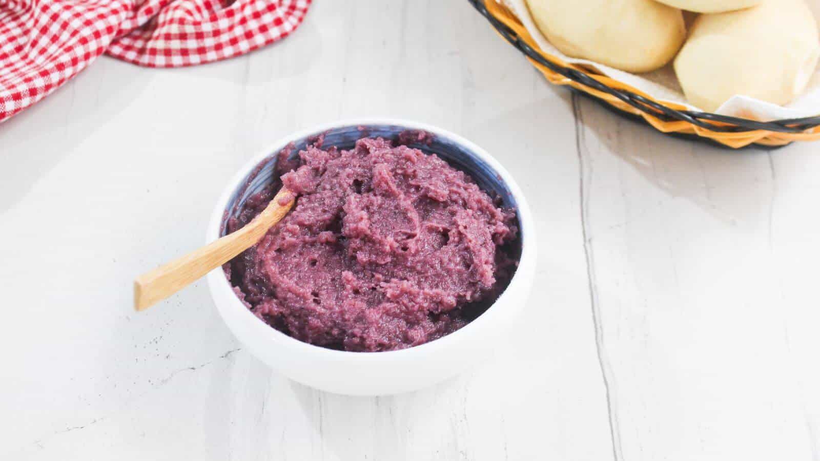 A white bowl filled with purple yam jam and a wooden spoon sits on a white surface near a basket of bread rolls.