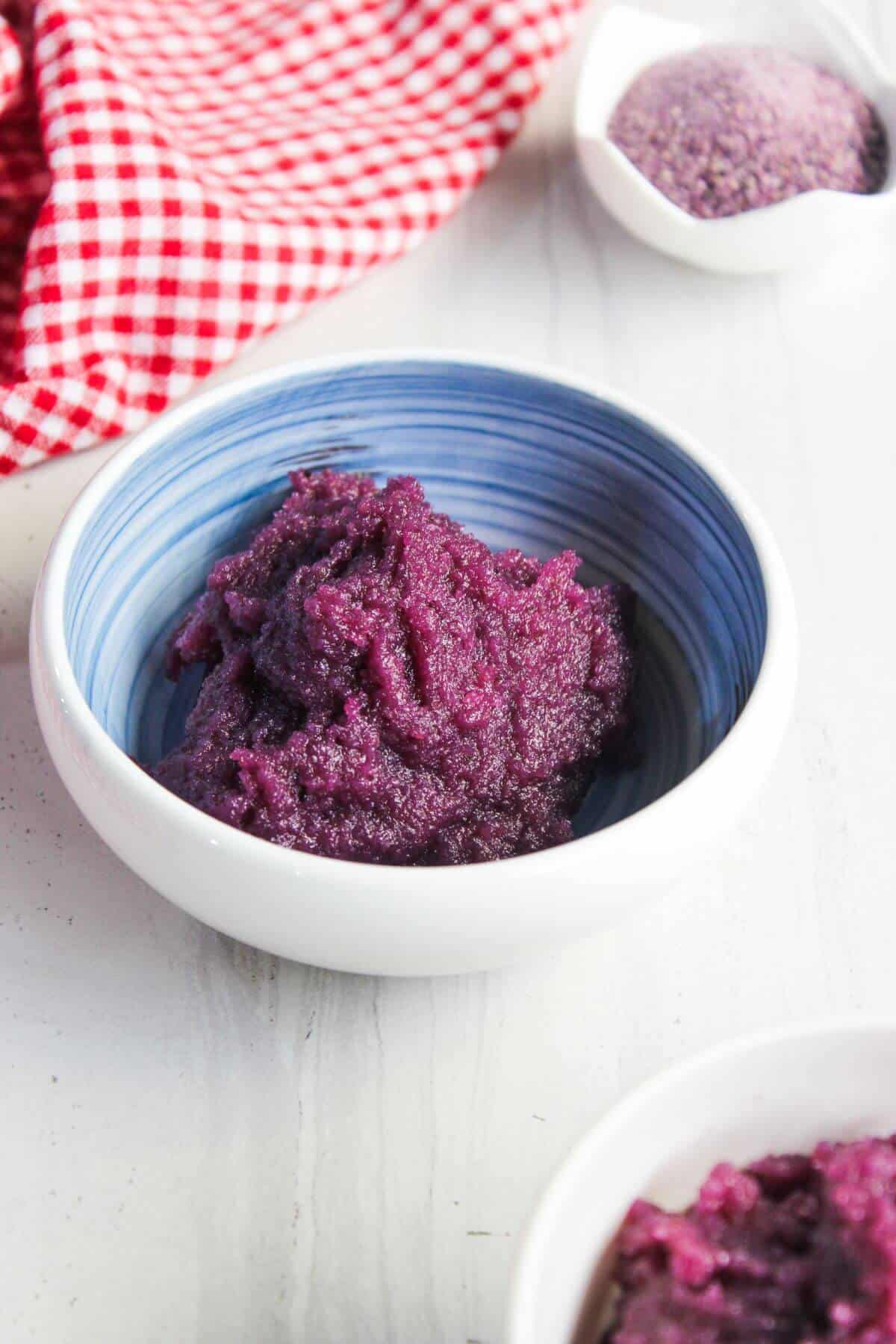 A bowl of purple yam paste sits on a white surface, with a red and white checkered cloth and a small bowl of purple powder in the background.