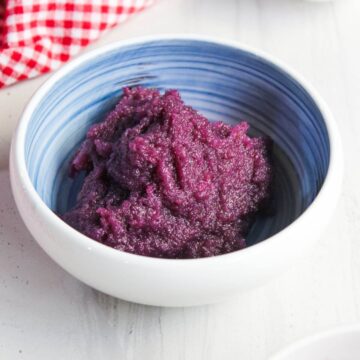 A bowl filled with purple ube halaya, a Filipino dessert made from purple yam, placed on a white surface with a red and white checked cloth partially visible.