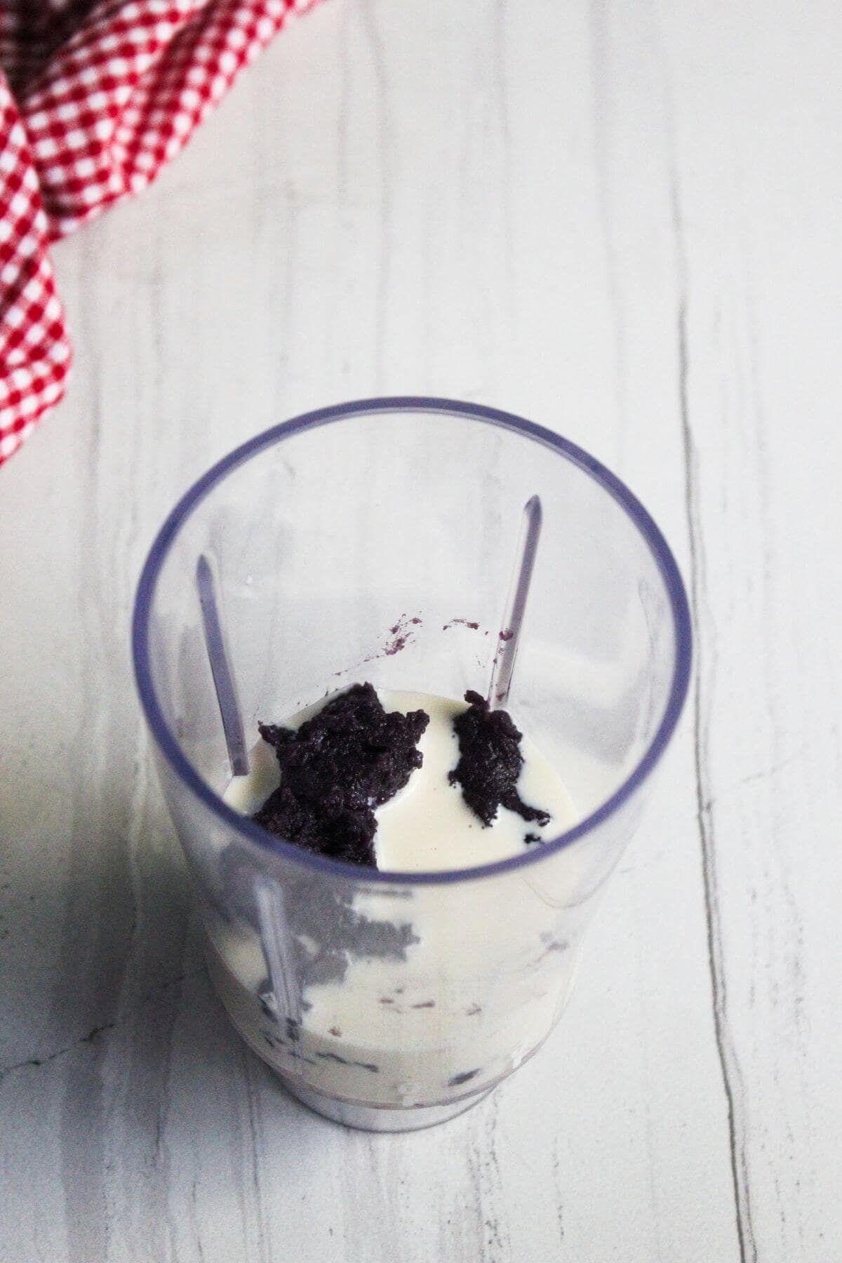 A blender cup containing white liquid and chunks of dark purple paste, placed on a white marbled surface next to a red and white checkered cloth.