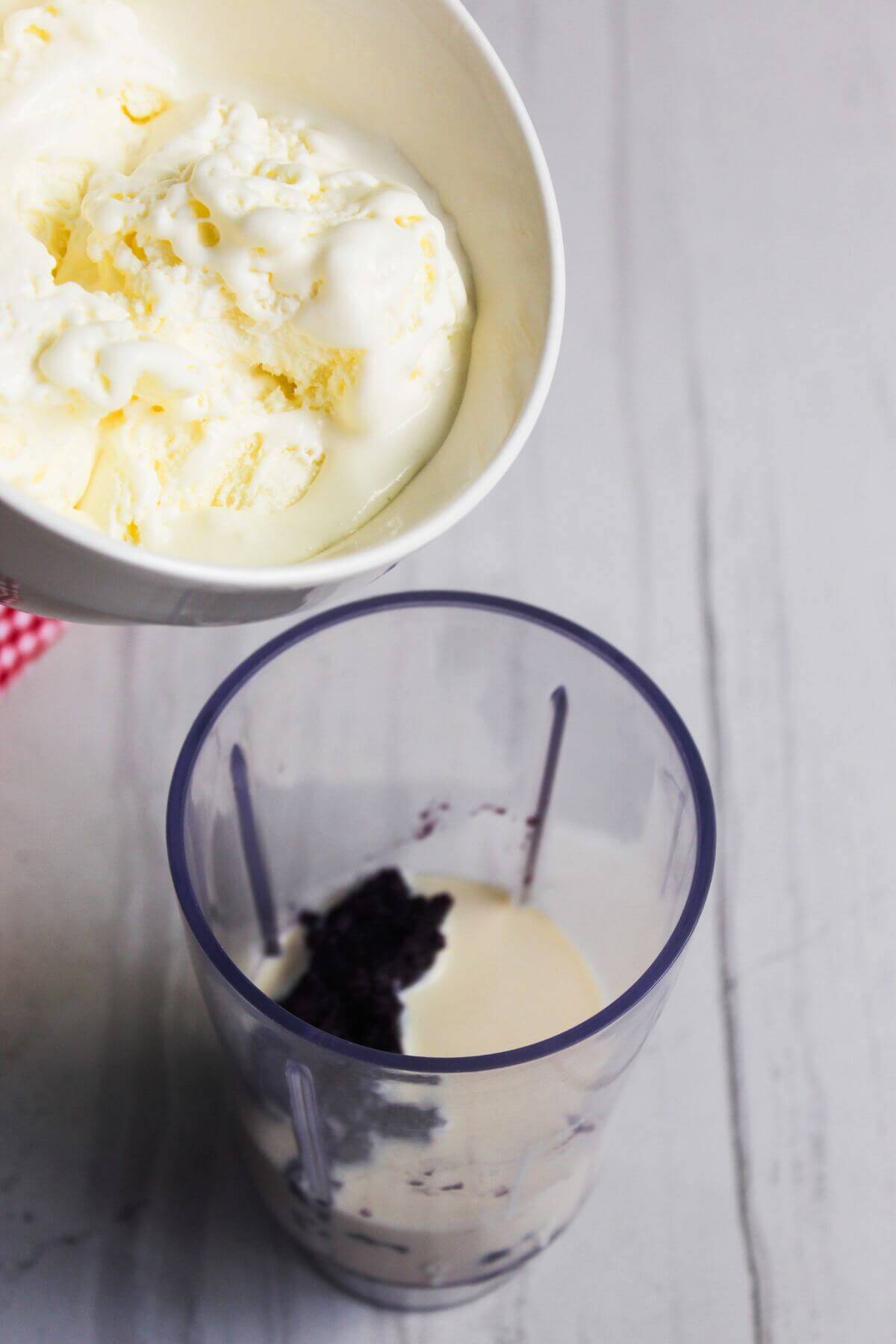 A bowl of vanilla ice cream is being held above a blender cup containing cream and blueberries on a light-colored surface.