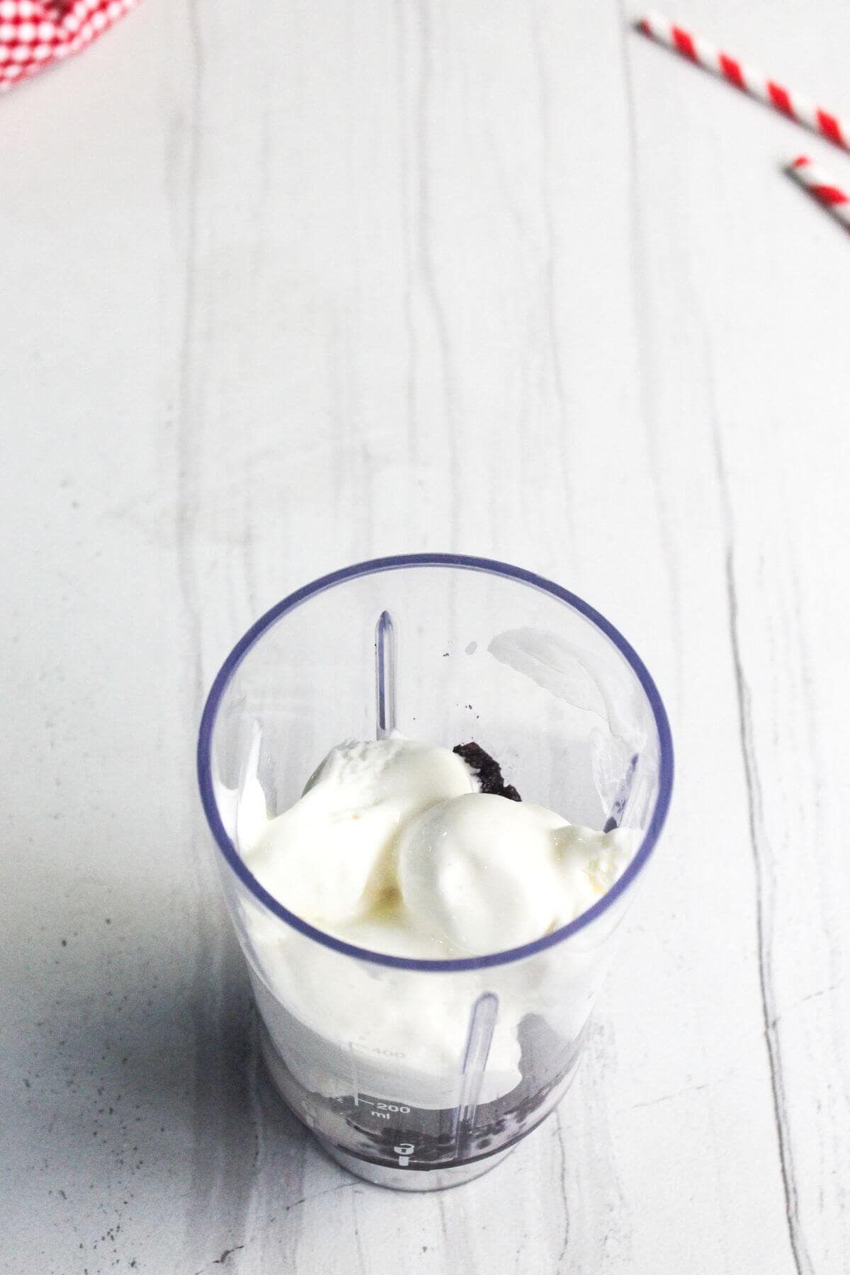 A blender cup containing ice cream and cookie pieces is placed on a white surface. Red-striped straws are visible in the background.
