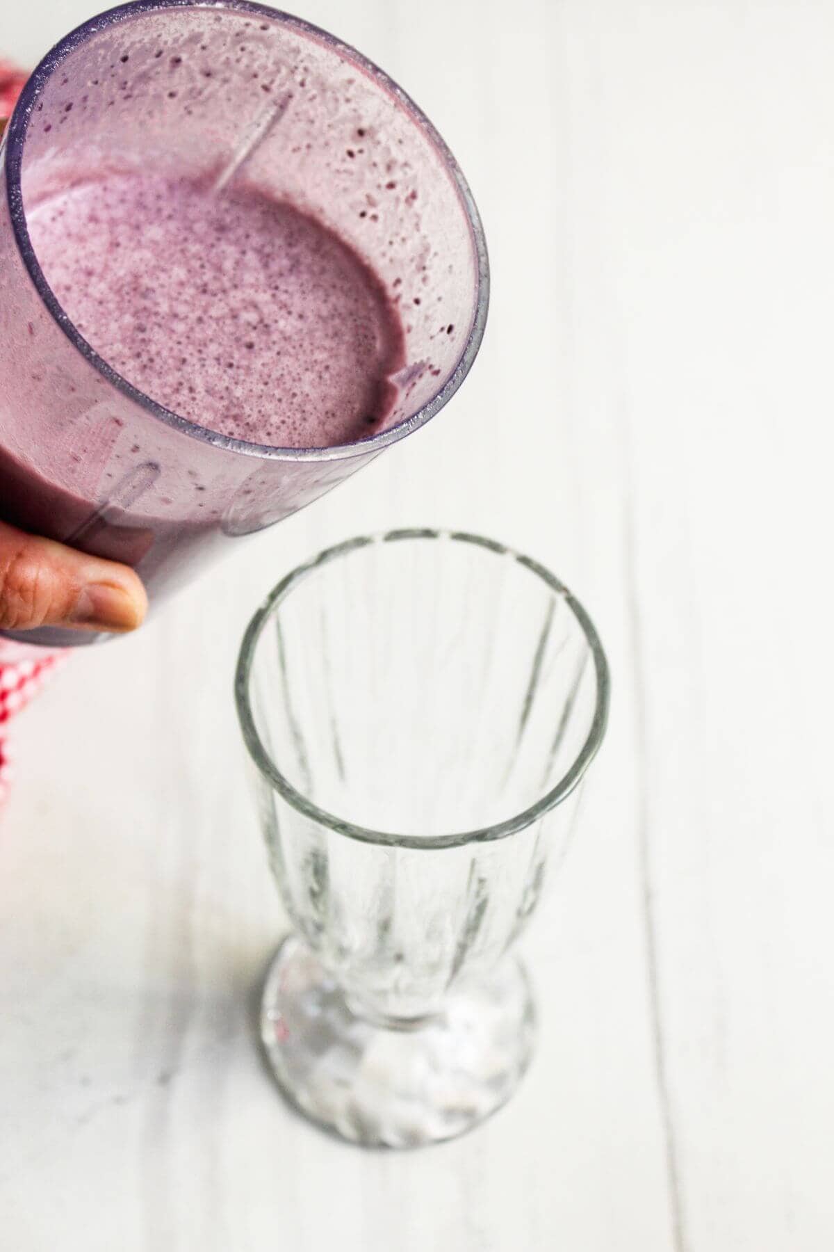 A hand holds a blender cup with a purple smoothie, positioned above an empty clear glass on a white surface.