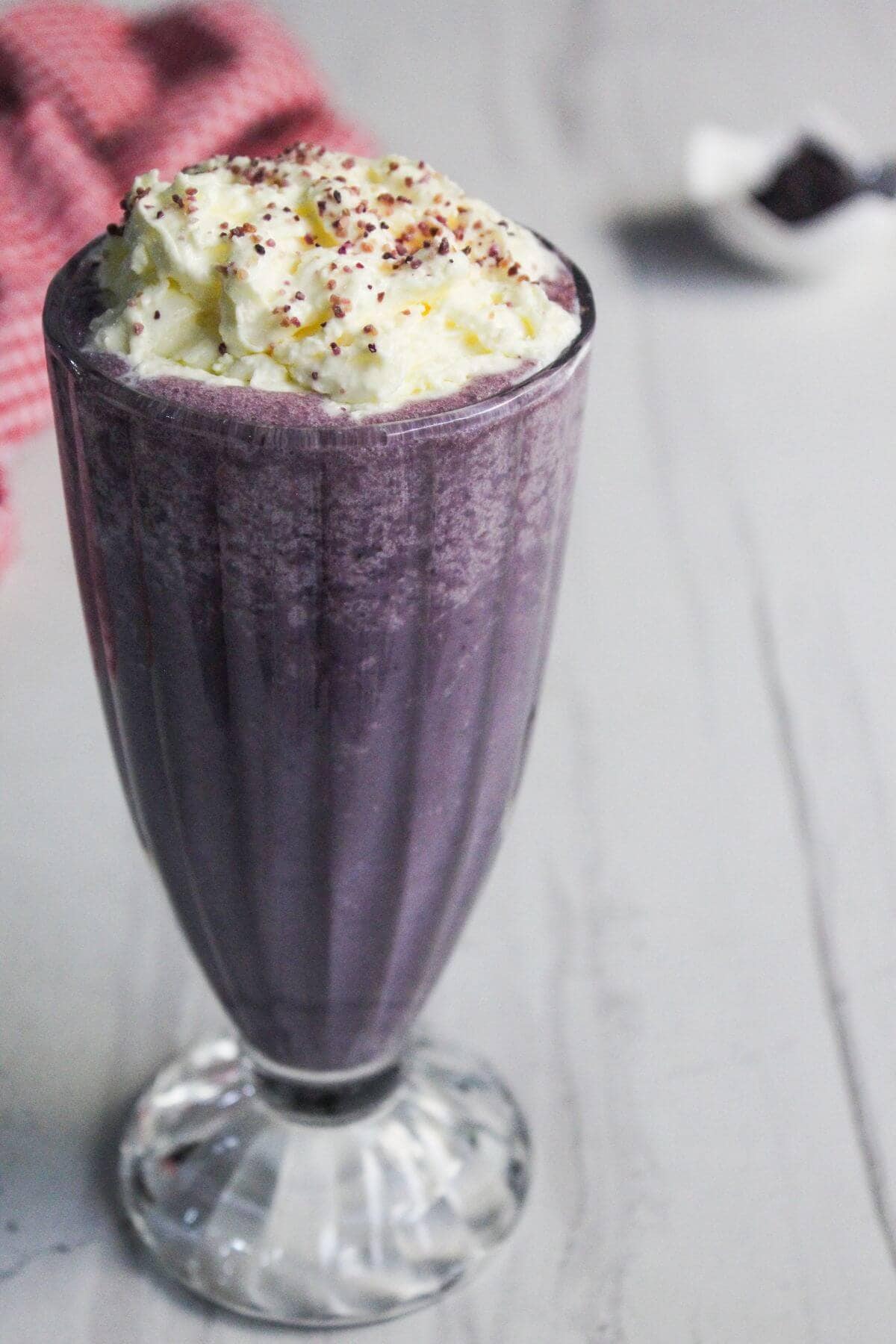 A tall glass of purple smoothie topped with whipped cream and sprinkles sits on a light gray surface.