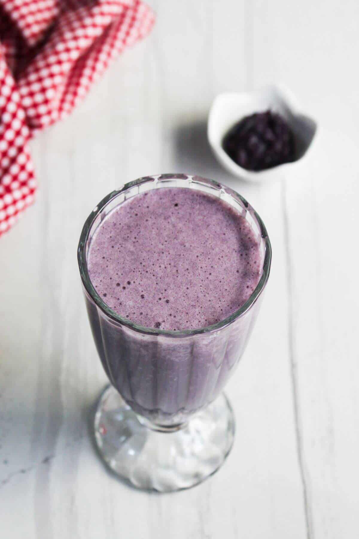 A glass filled with a purple smoothie sits on a white surface, with a small bowl of dark fruit in the background and a red-checkered cloth to the side.