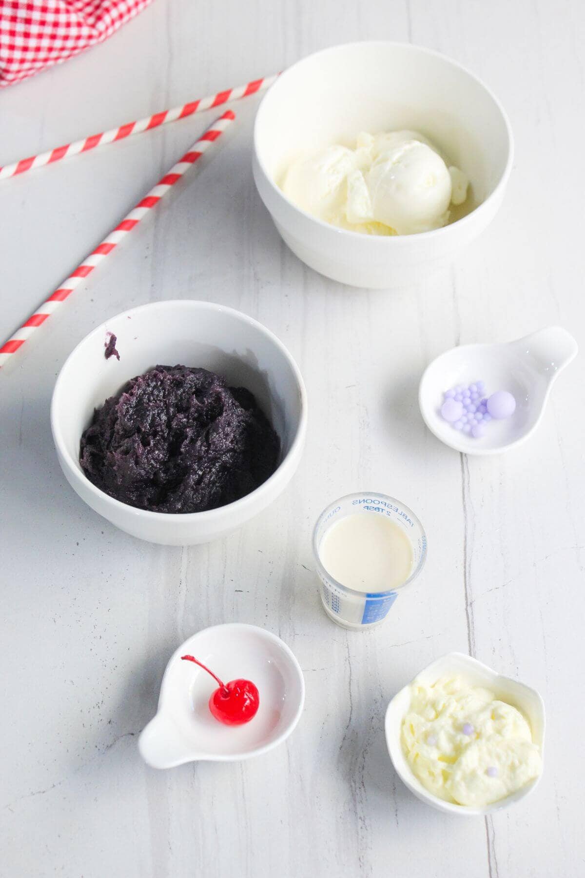 Bowls containing ice cream, purple yam paste, a cherry, evaporated milk, and toppings are arranged on a white surface with two red-striped straws in the background.