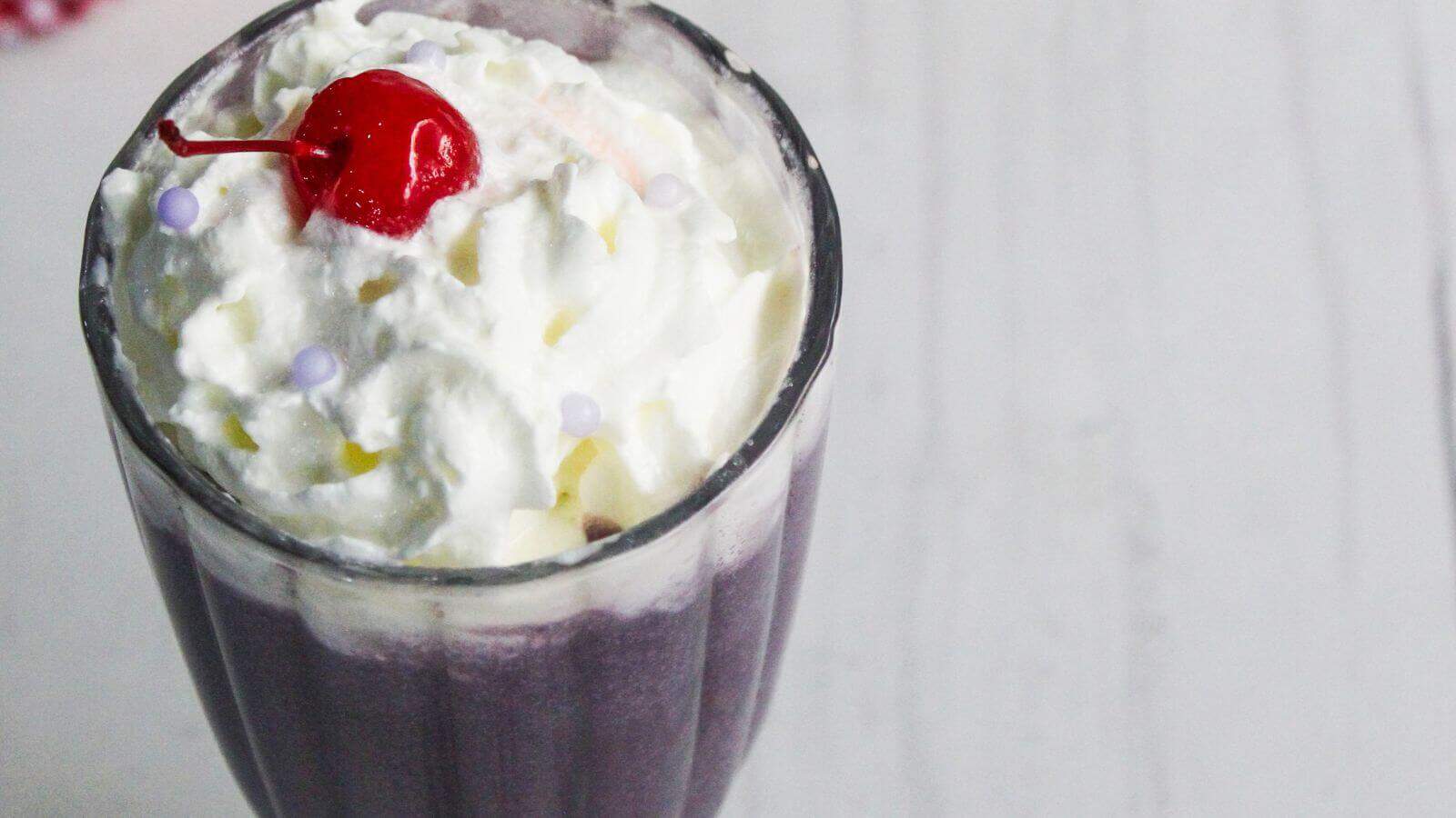 A glass of purple milkshake topped with whipped cream, a red cherry, and small white candy pearls, set on a light-colored surface.