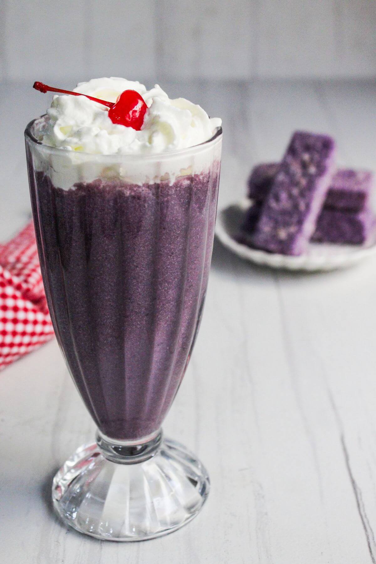 A tall glass of purple milkshake topped with whipped cream and a cherry, with a plate of purple snacks in the background on a white surface.