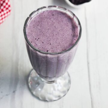 A clear glass filled with a thick, purple smoothie sits on a white surface.