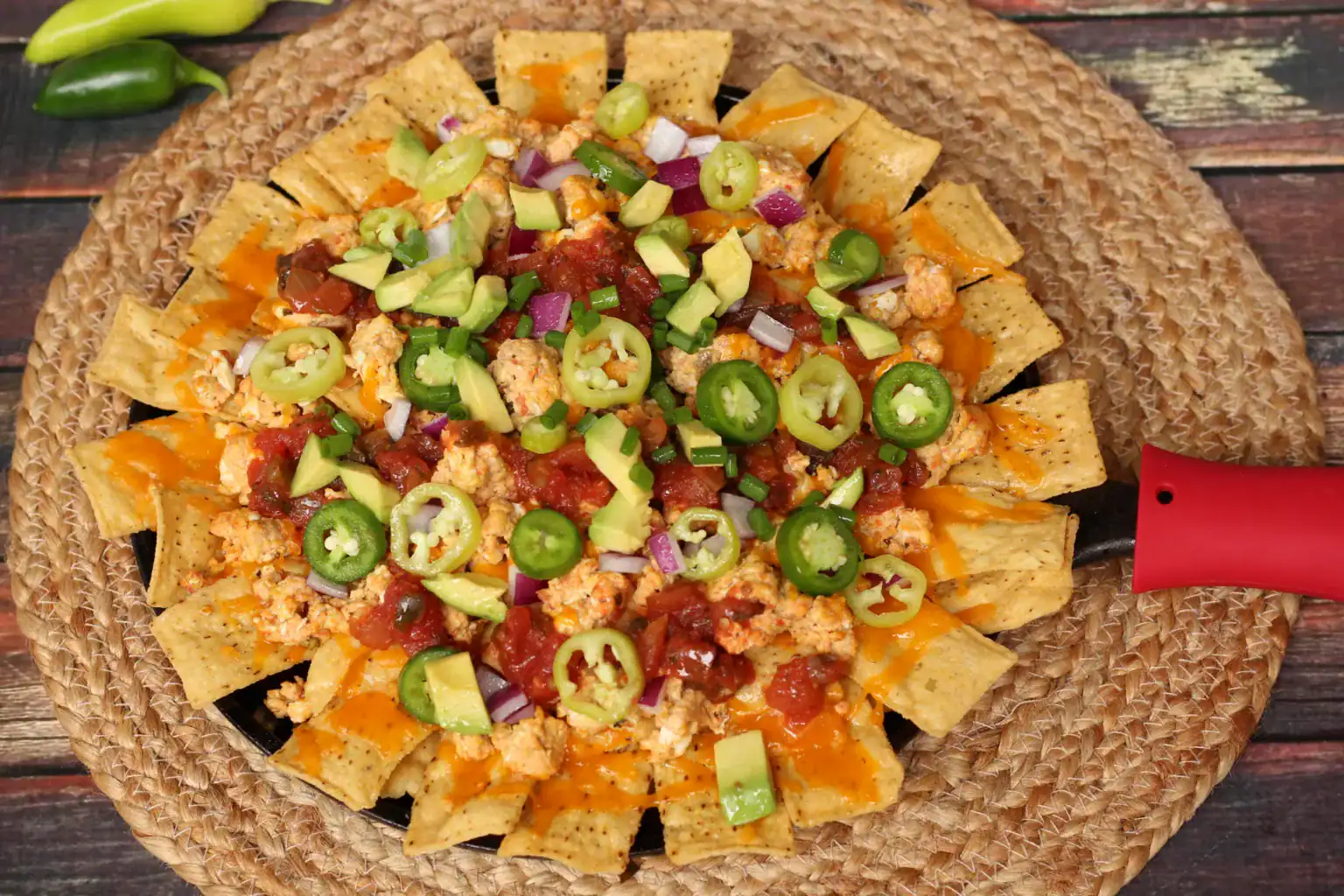 A plate of nachos topped with melted cheese, diced avocado, jalapeño slices, red onion, salsa, and ground meat on a woven placemat.