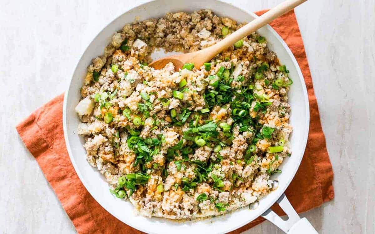 A skillet filled with cooked ground meat, chopped green onions, herbs, and cauliflower rice sits on an orange cloth with a wooden spoon.