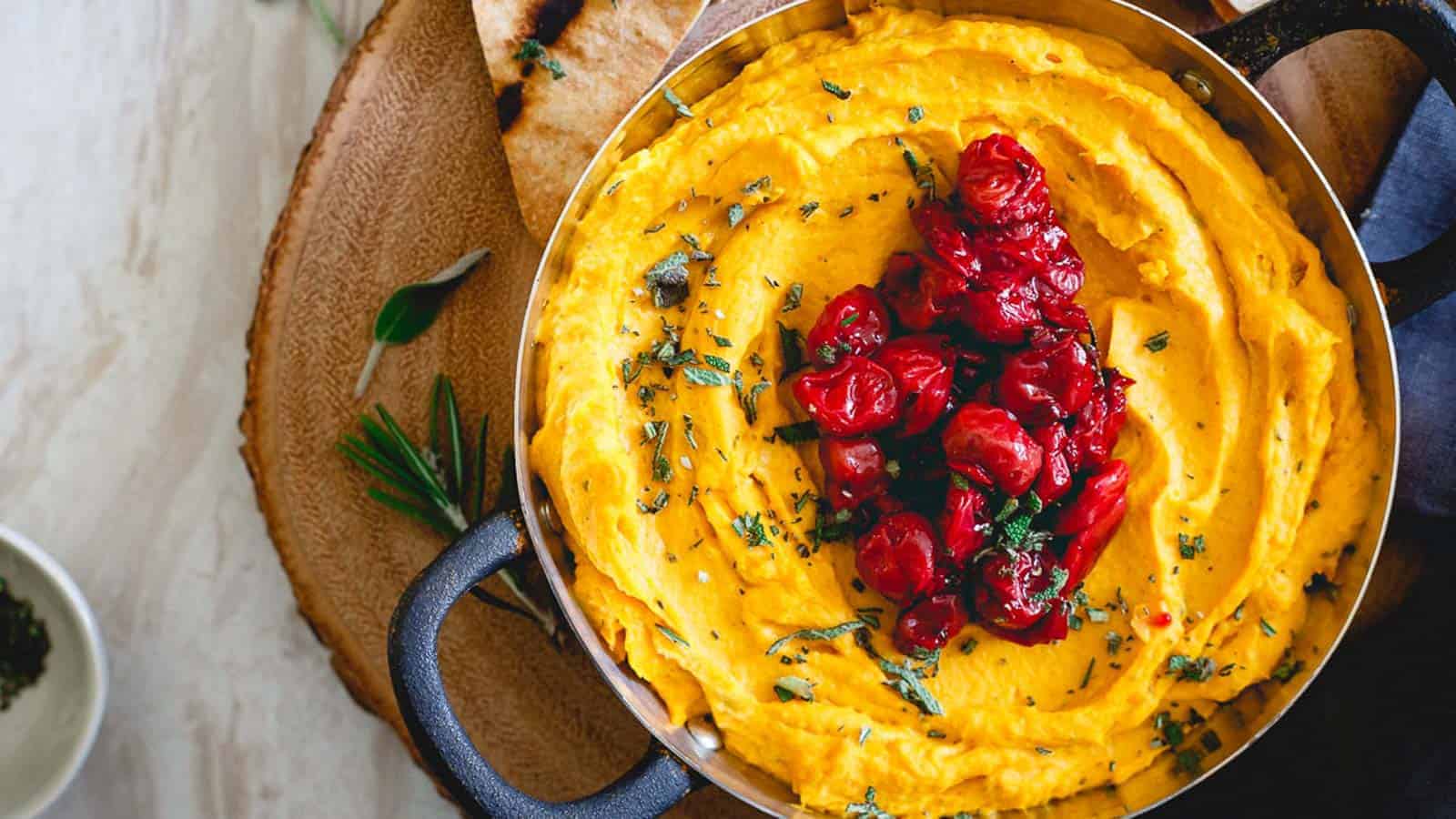 A pot of creamy mashed sweet potatoes topped with roasted cherry tomatoes and chopped herbs, placed on a wooden board with flatbread on the side.