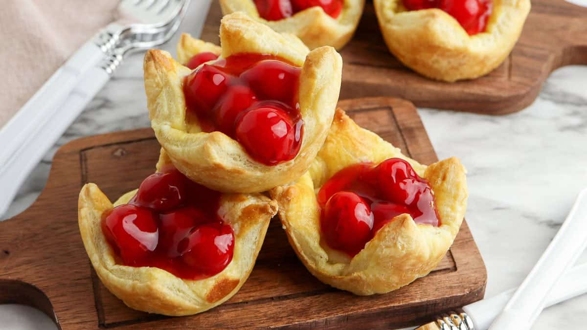 Three cherry puff pastry tarts with bright red cherry filling stacked on a wooden serving board, with silverware visible in the background.