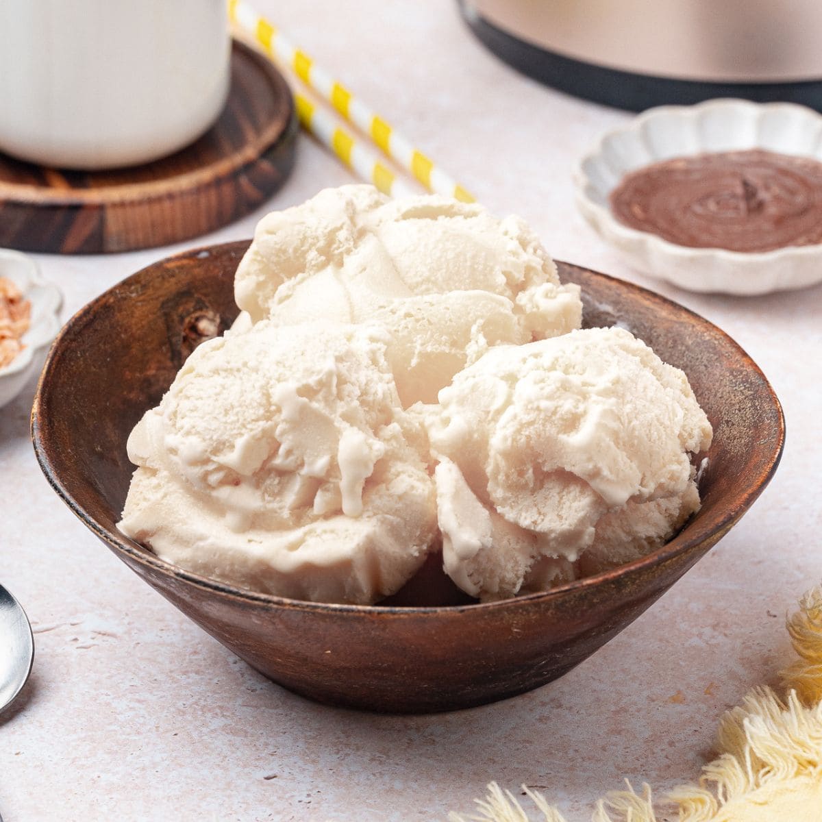 A brown bowl filled with several scoops of coconut milk ice cream sits on a light surface, with a spoon and other dishes visible in the background.