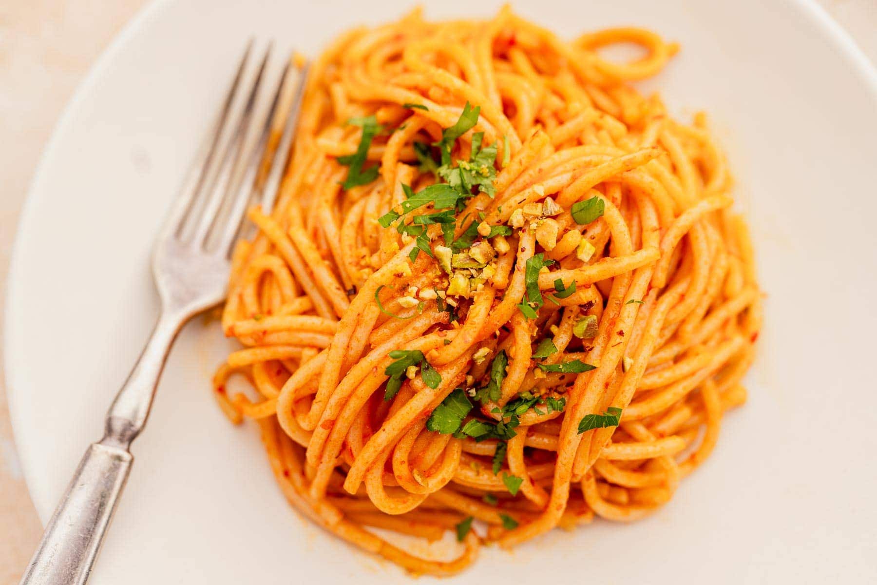 A plate of spaghetti topped with red sauce, chopped herbs, and crushed nuts, with a fork placed beside the pasta.
