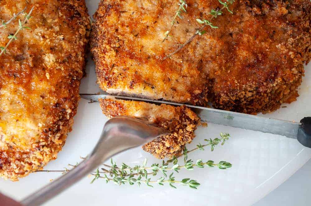 A breaded and baked chicken breast being sliced with a knife and fork on a white plate, garnished with fresh thyme.