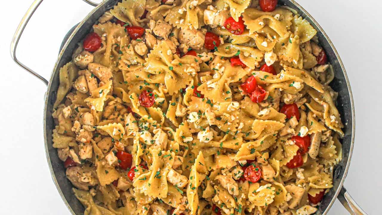A pan filled with bowtie pasta, diced chicken, cherry tomatoes, and crumbled cheese, garnished with chopped herbs.