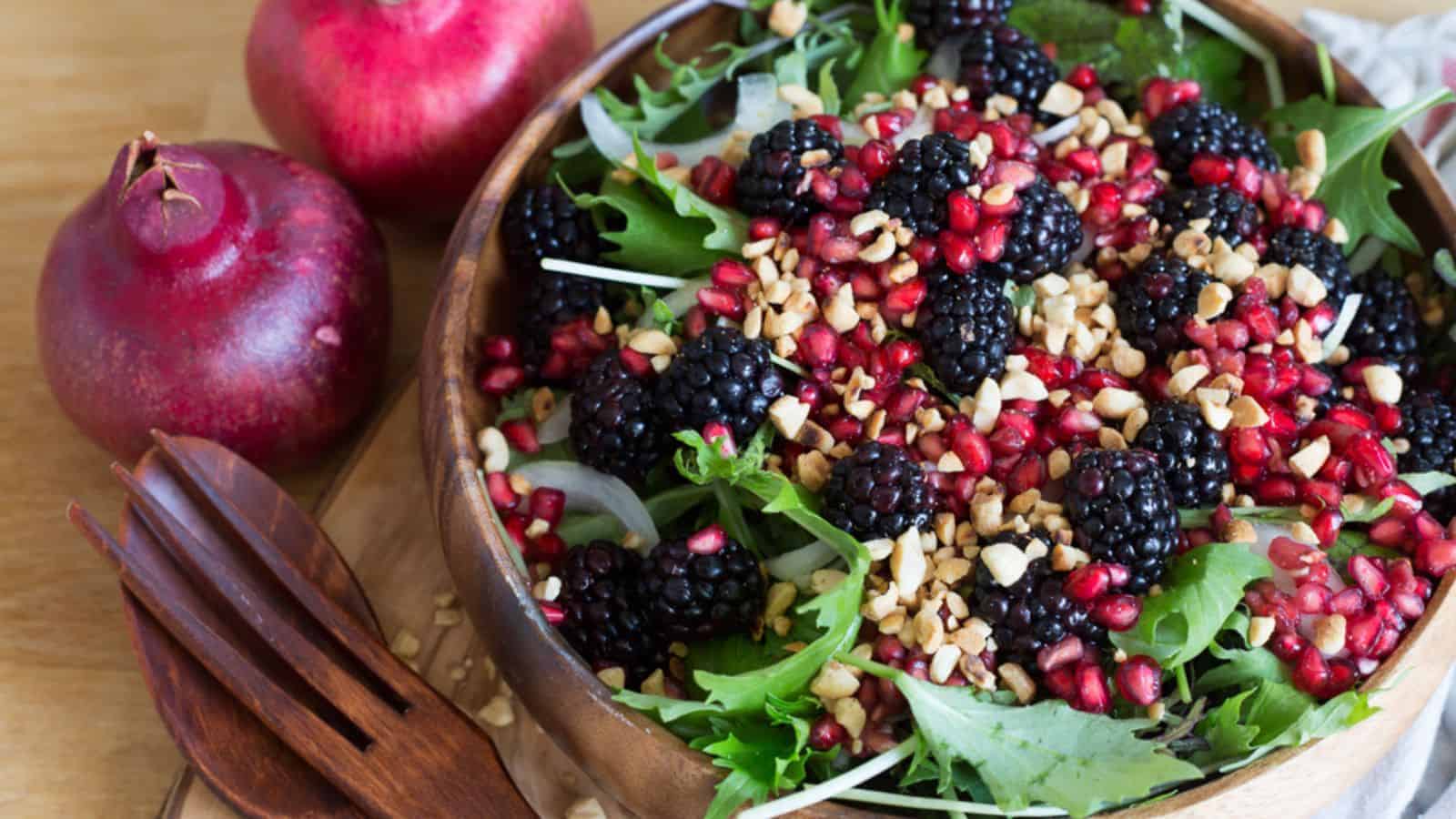 A wooden bowl of salad with mixed greens, blackberries, pomegranate seeds, and chopped nuts, next to two whole pomegranates and wooden salad utensils.