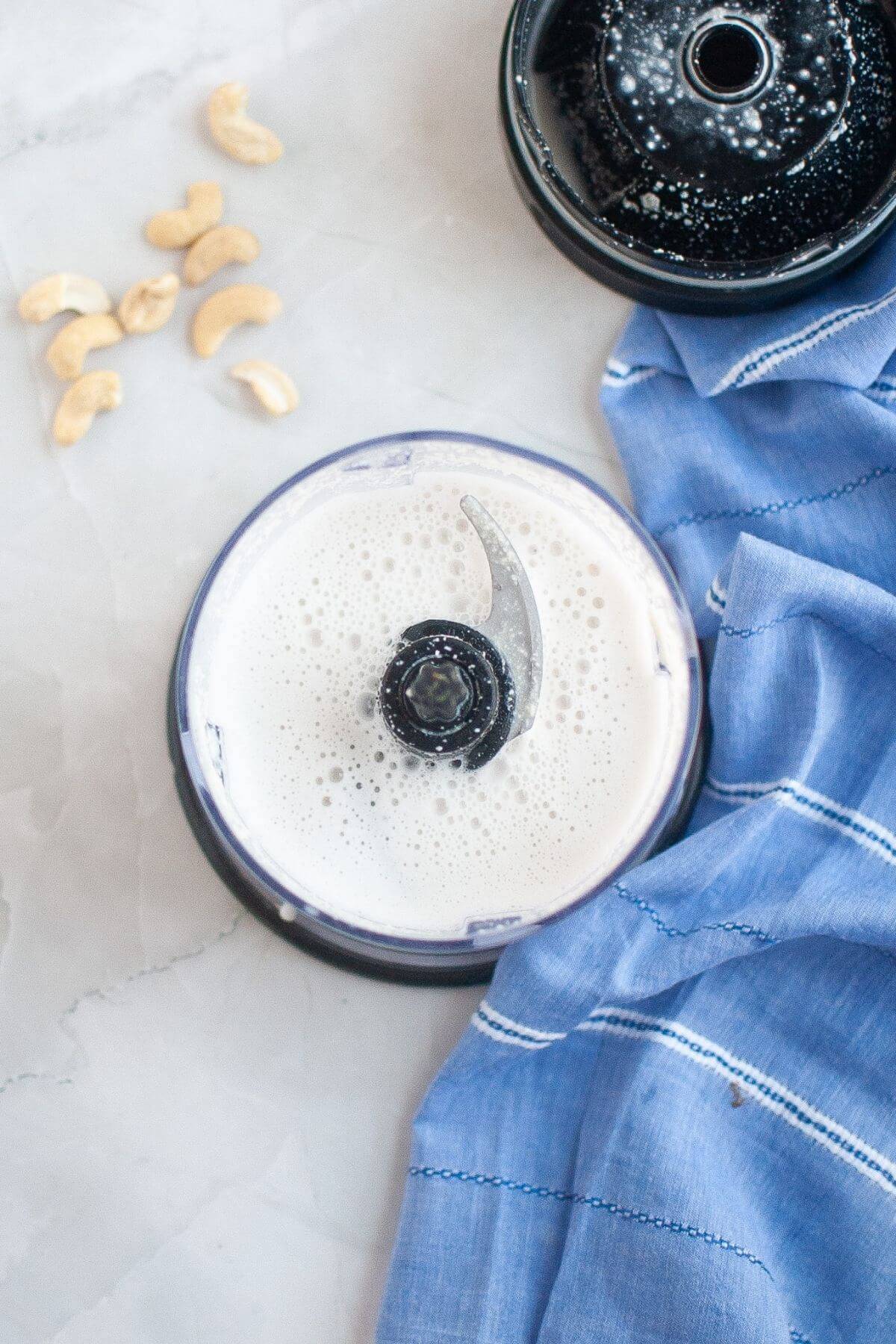 A blender with blended cashew milk inside sits on a marble surface next to a blue cloth and some whole cashews.