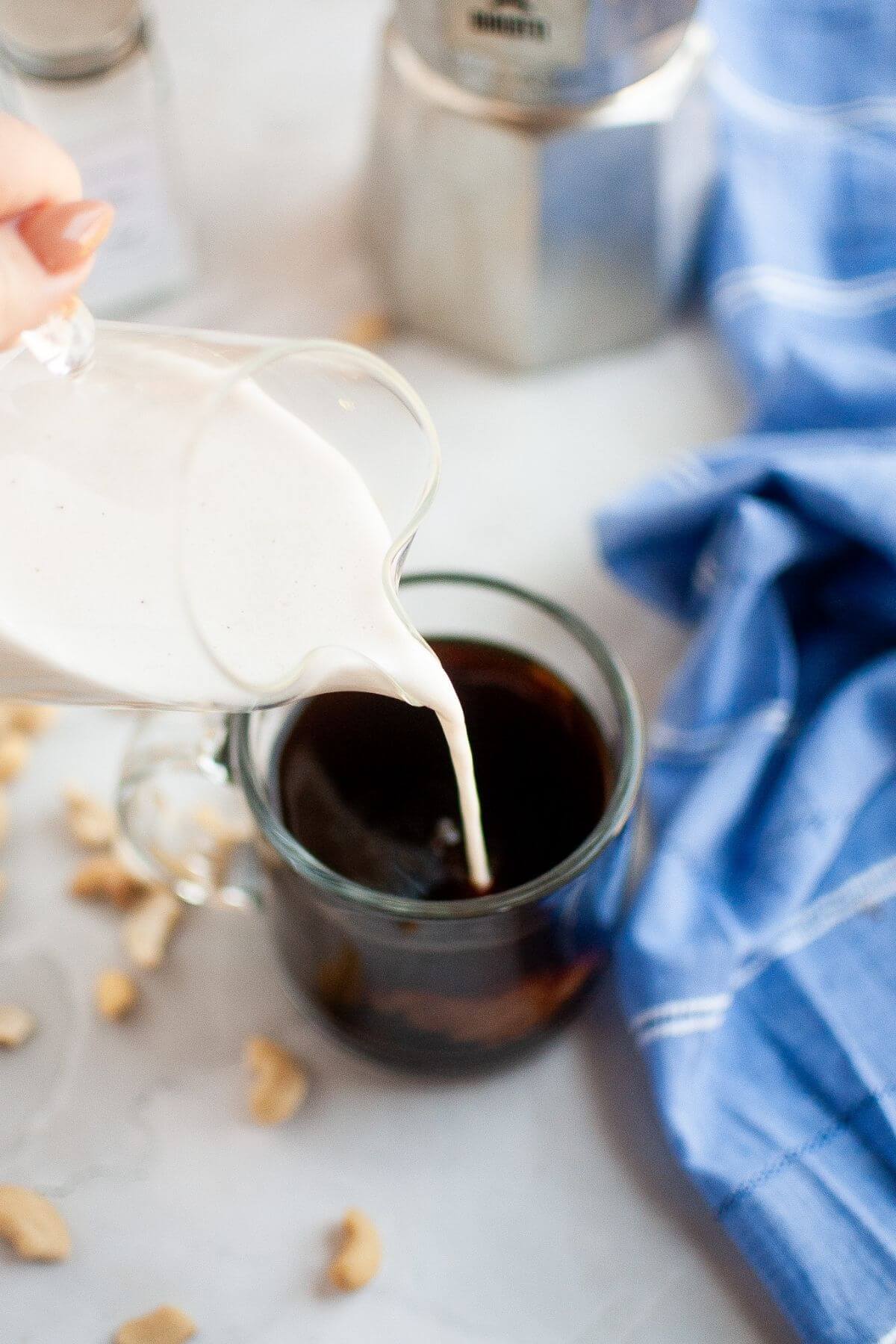 A hand pours creamy liquid from a small glass pitcher into a cup of black coffee, with a blue cloth and metal coffee maker in the background.