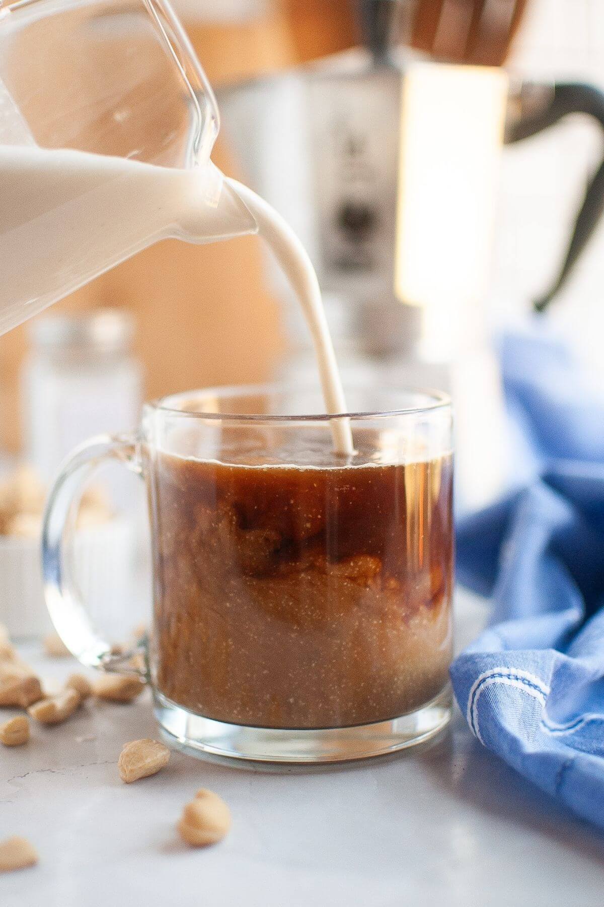 A clear glass mug of iced coffee is being filled with milk from a glass pitcher, with some coffee beans and a blue cloth nearby.