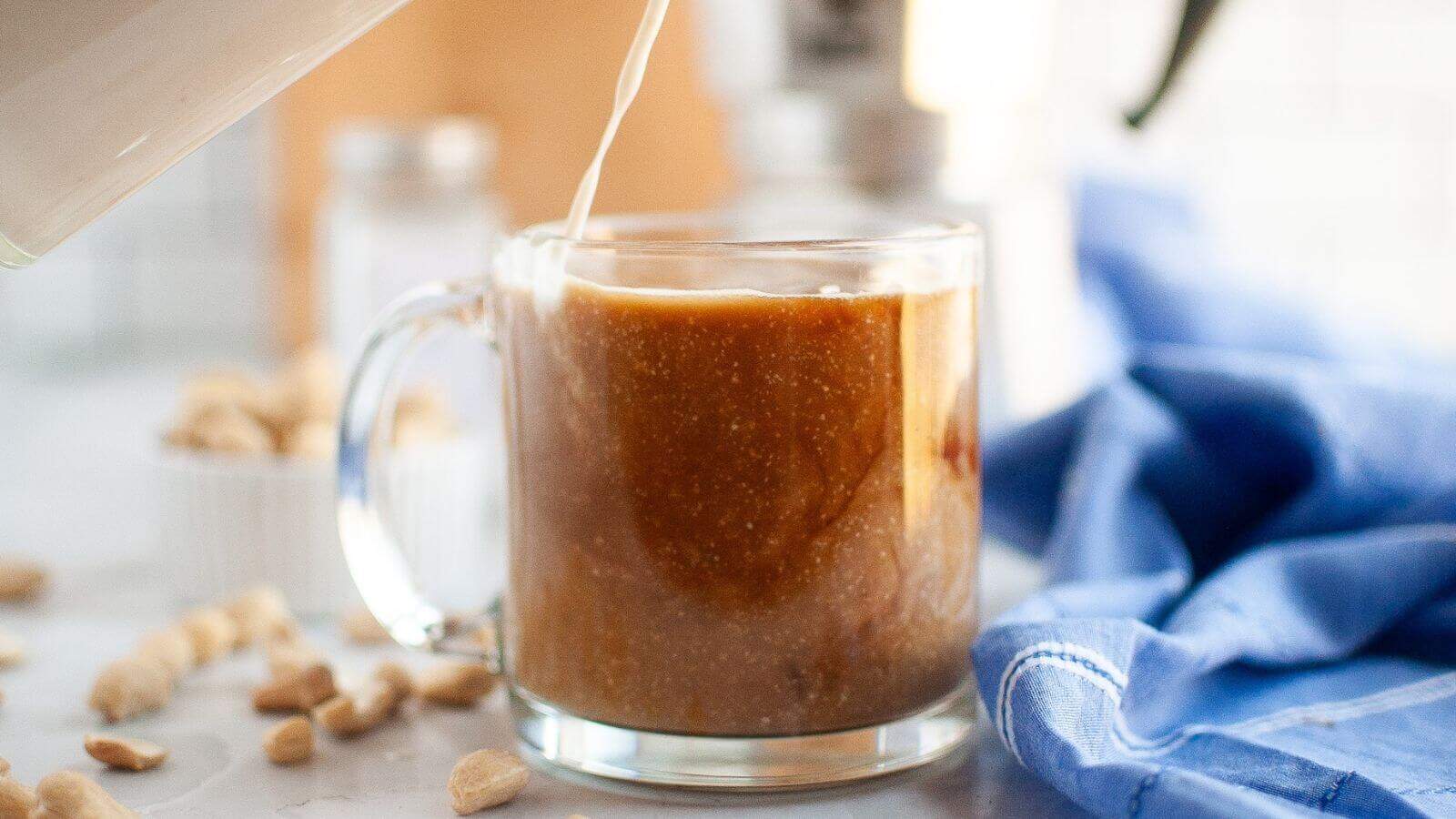 A glass mug of coffee with milk being poured in, surrounded by peanuts and a blue cloth on a light countertop.