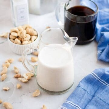 A glass pitcher of cashew milk sits on a counter near a bowl of cashews, a glass mug of coffee, and a blue cloth.