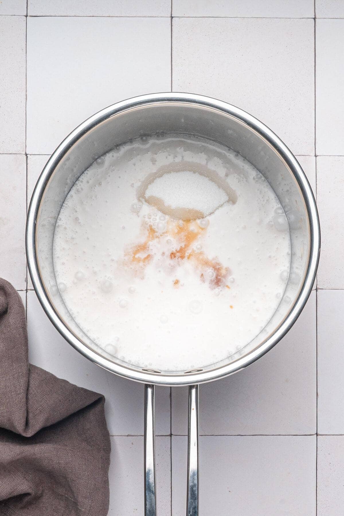 A saucepan filled with milk, sugar, and vanilla extract sits on a white tiled surface next to a brown cloth.