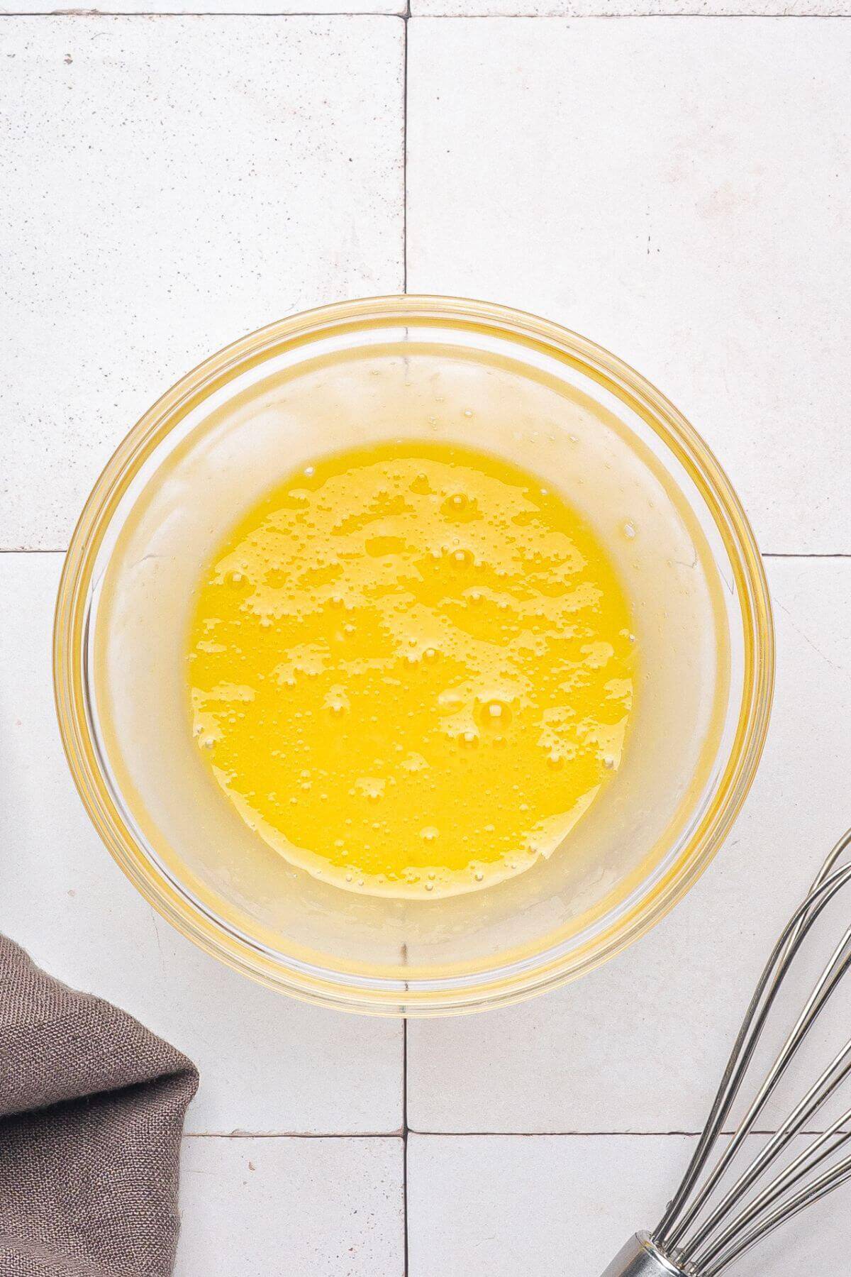 A glass bowl containing melted butter sits on a tiled surface next to a metal whisk and a brown cloth.