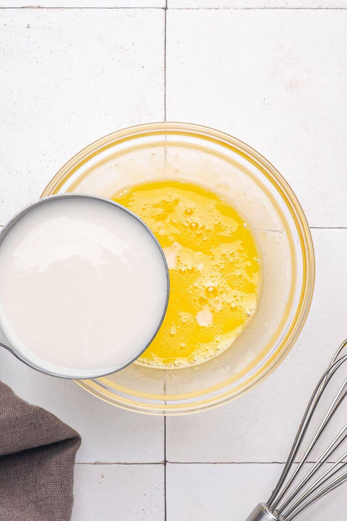 A bowl of whisked eggs with milk being poured in, next to a metal whisk on a white tiled surface.