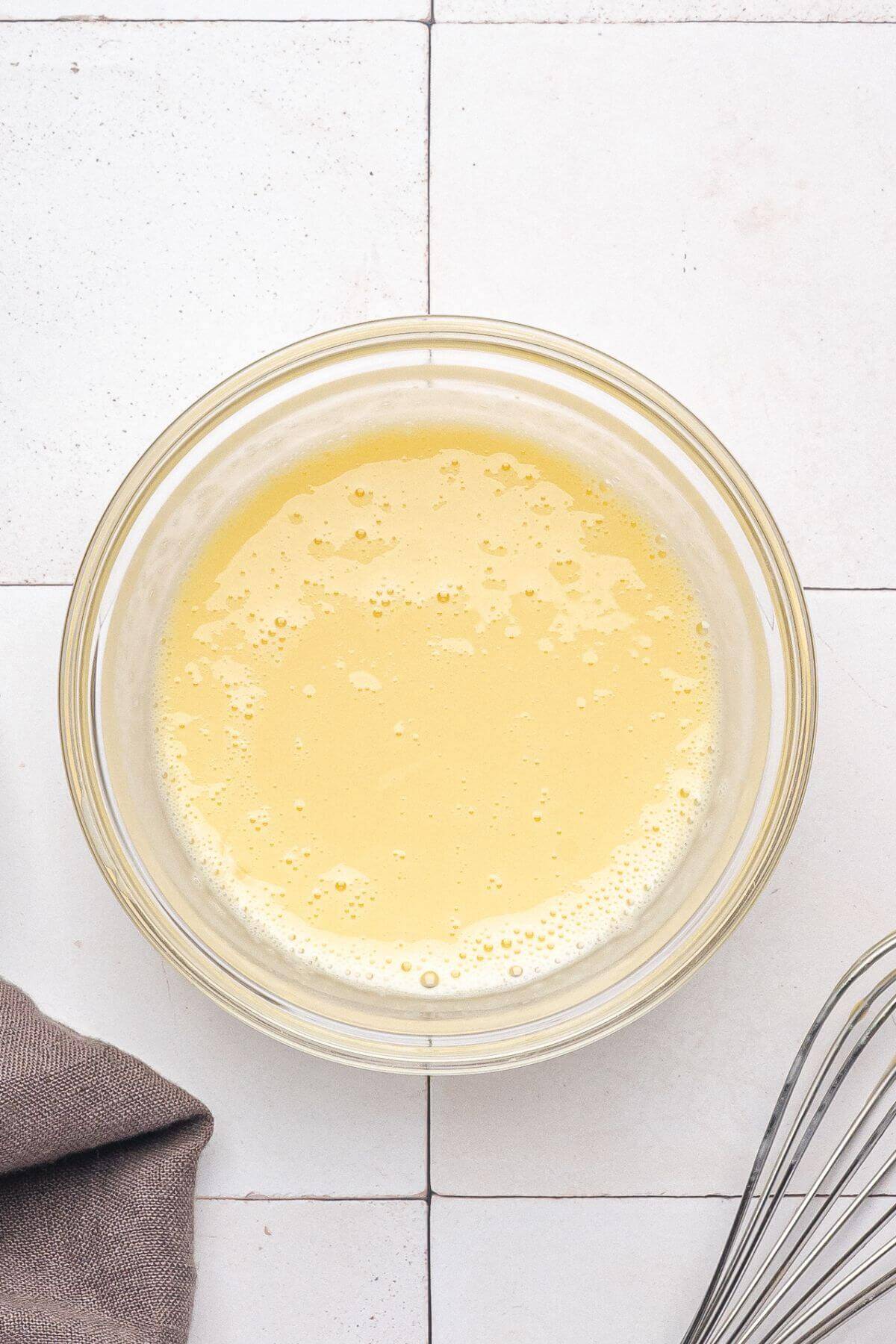 A glass bowl filled with a pale yellow liquid mixture on a tiled surface, with part of a whisk and a gray cloth visible nearby.