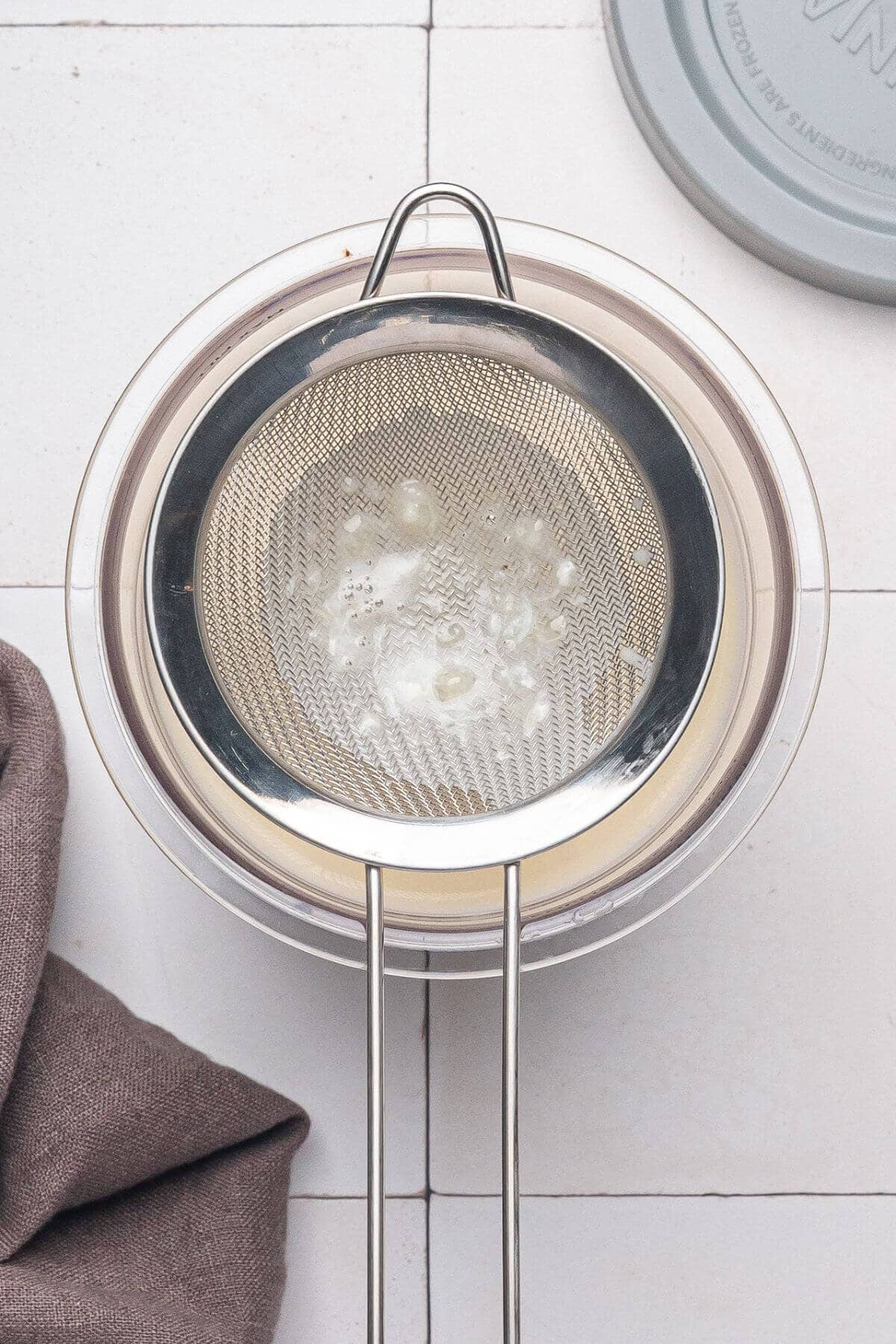 A metal strainer sits over a glass bowl with white liquid and curds inside, next to a gray cloth and a round lid on a tiled surface.
