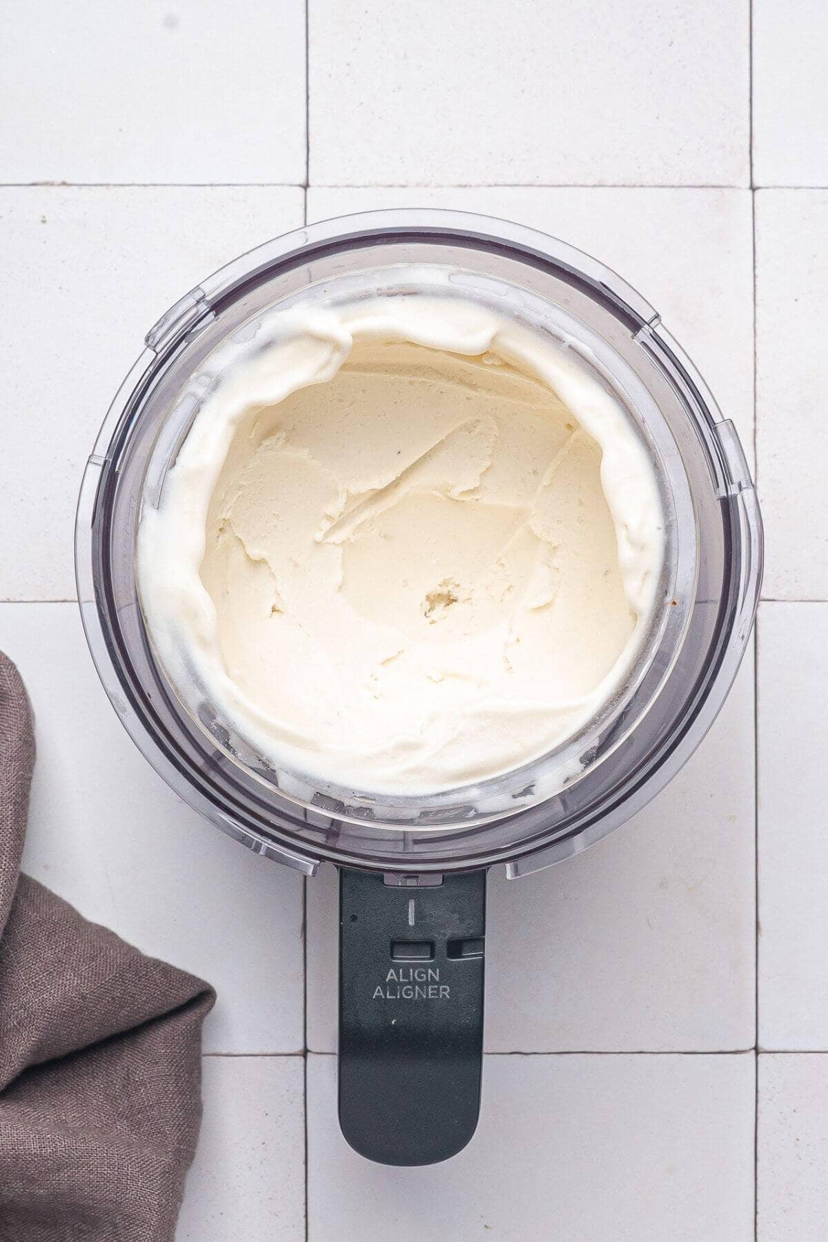 A food processor bowl filled with smooth, creamy vanilla ice cream sits on a white tiled surface next to a folded brown cloth.