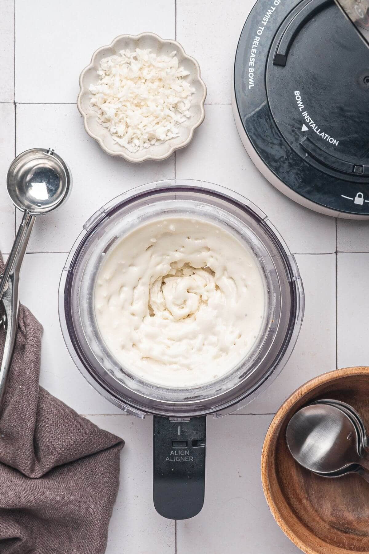 A food processor filled with a creamy white mixture sits on a tiled surface, surrounded by a scoop, a small dish of shredded coconut, a wooden bowl, and a cloth napkin.