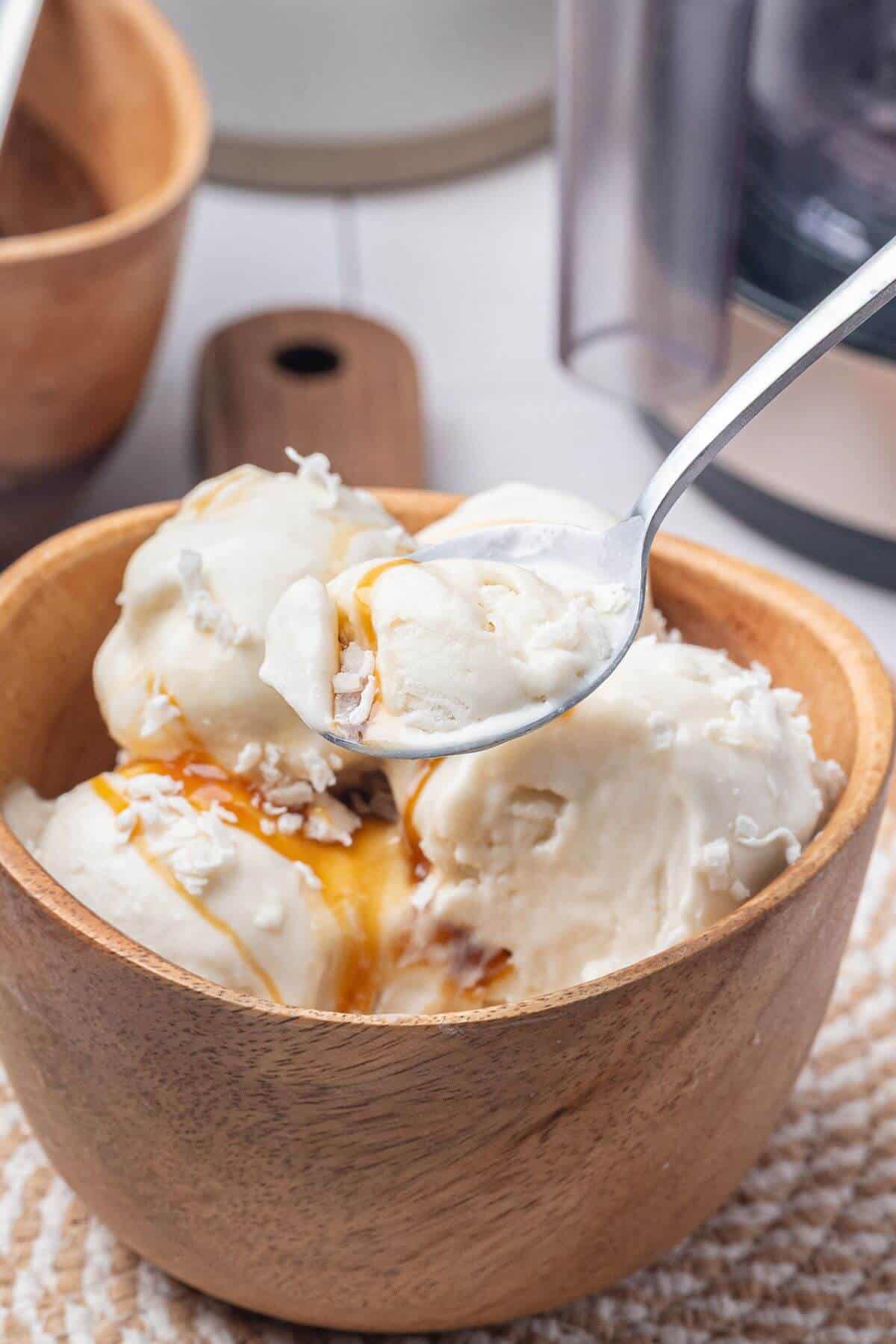 A wooden bowl filled with scoops of vanilla ice cream topped with caramel sauce and shredded coconut, with a spoon lifting a bite.