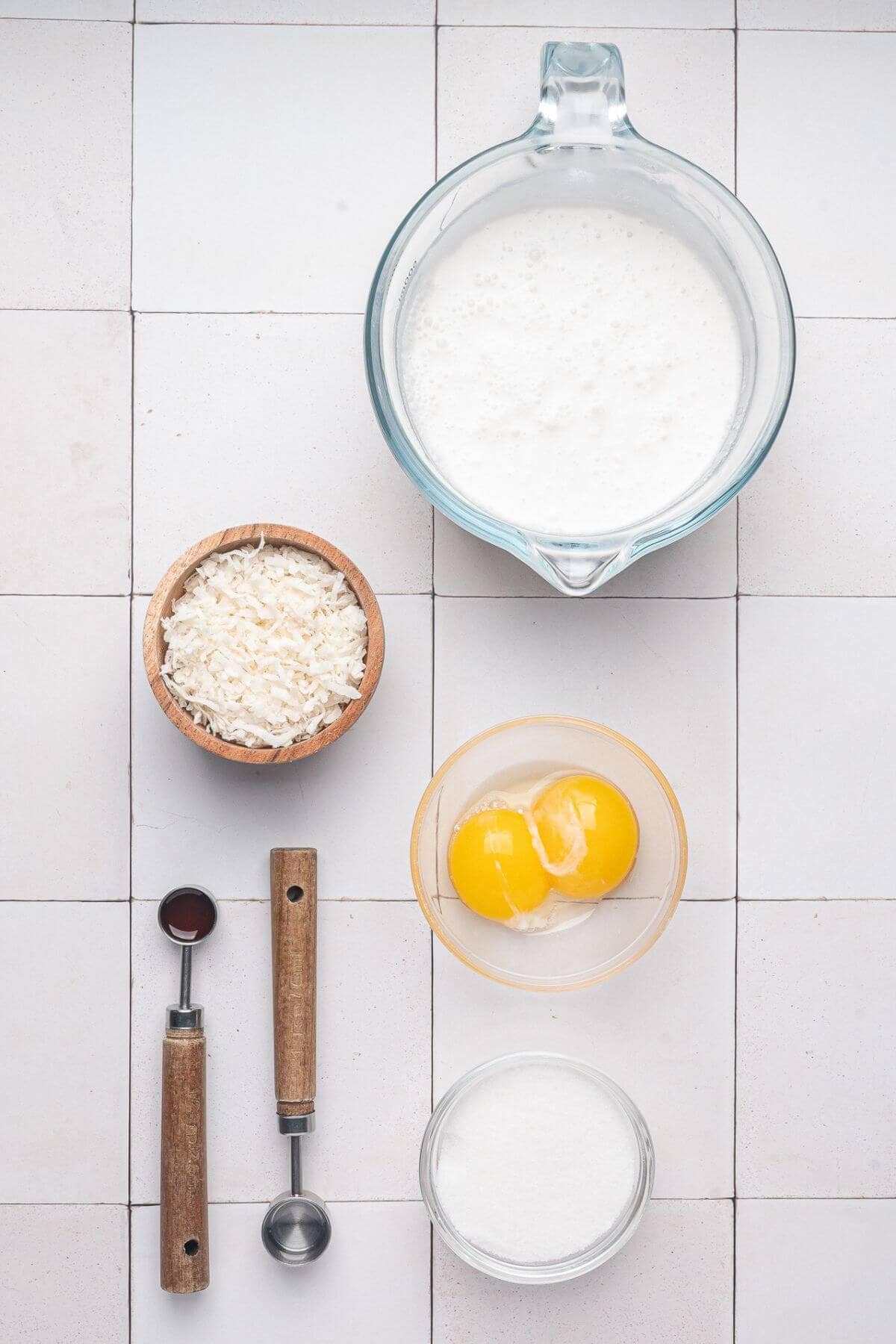 Measuring cup with milk, bowl of shredded coconut, two egg yolks in a bowl, sugar, vanilla extract, and a teaspoon on a tiled surface.
