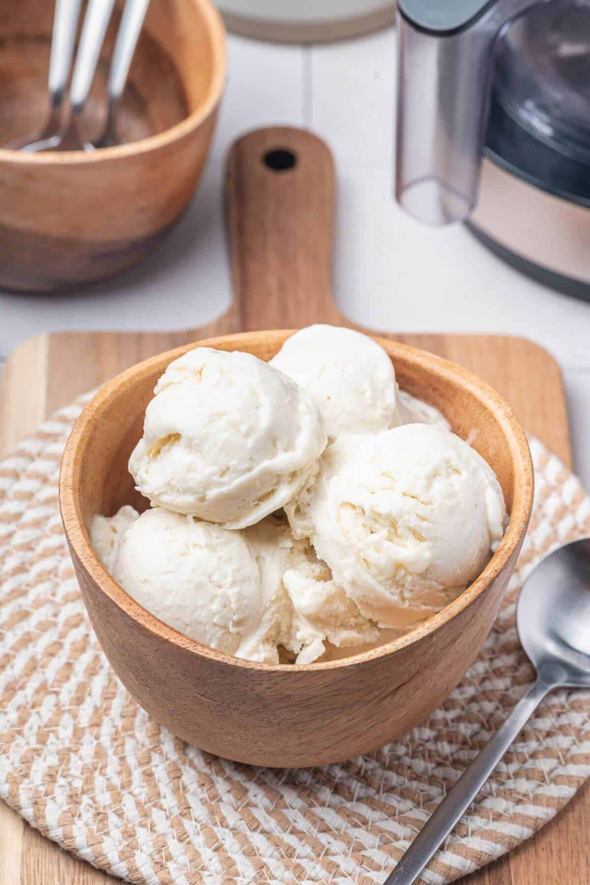 A wooden bowl filled with several scoops of vanilla ice cream sits on a woven mat beside a metal spoon.