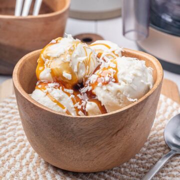 A wooden bowl filled with three scoops of vanilla ice cream, topped with caramel sauce and shredded coconut, sits on a woven placemat next to a metal spoon.