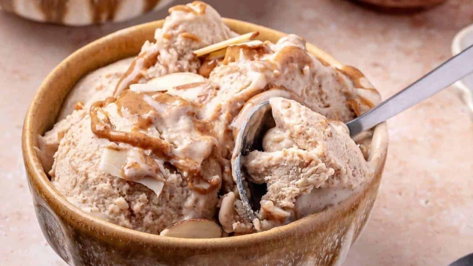 A bowl of light brown ice cream with a spoon, topped with sliced almonds and a light drizzle of syrup.