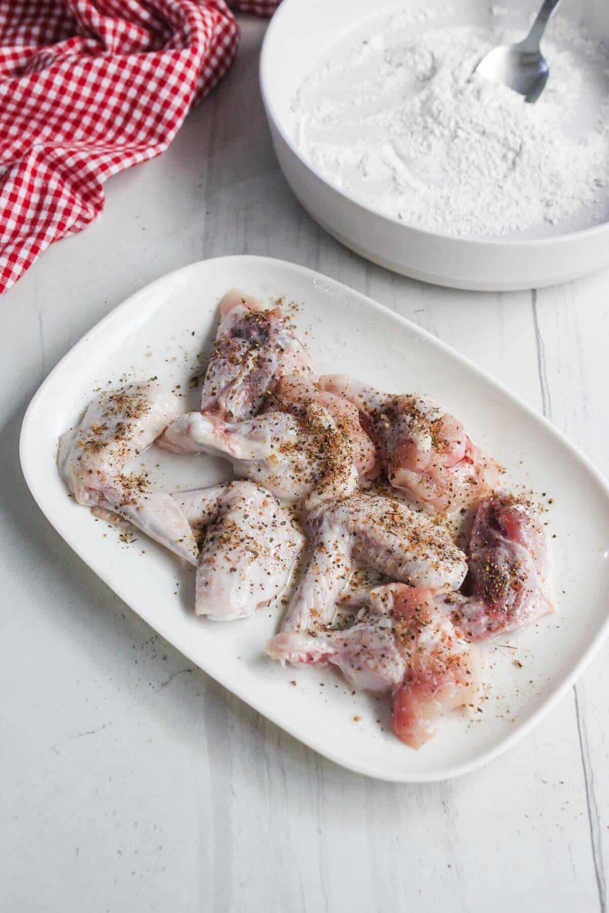 Raw chicken wings seasoned with spices on a white plate, with a bowl of flour and a spoon in the background on a white surface.