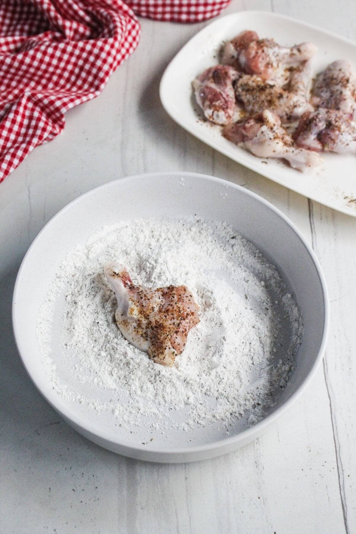 A piece of seasoned raw chicken wing is being coated with flour in a white bowl, with more floured chicken pieces on a plate nearby.