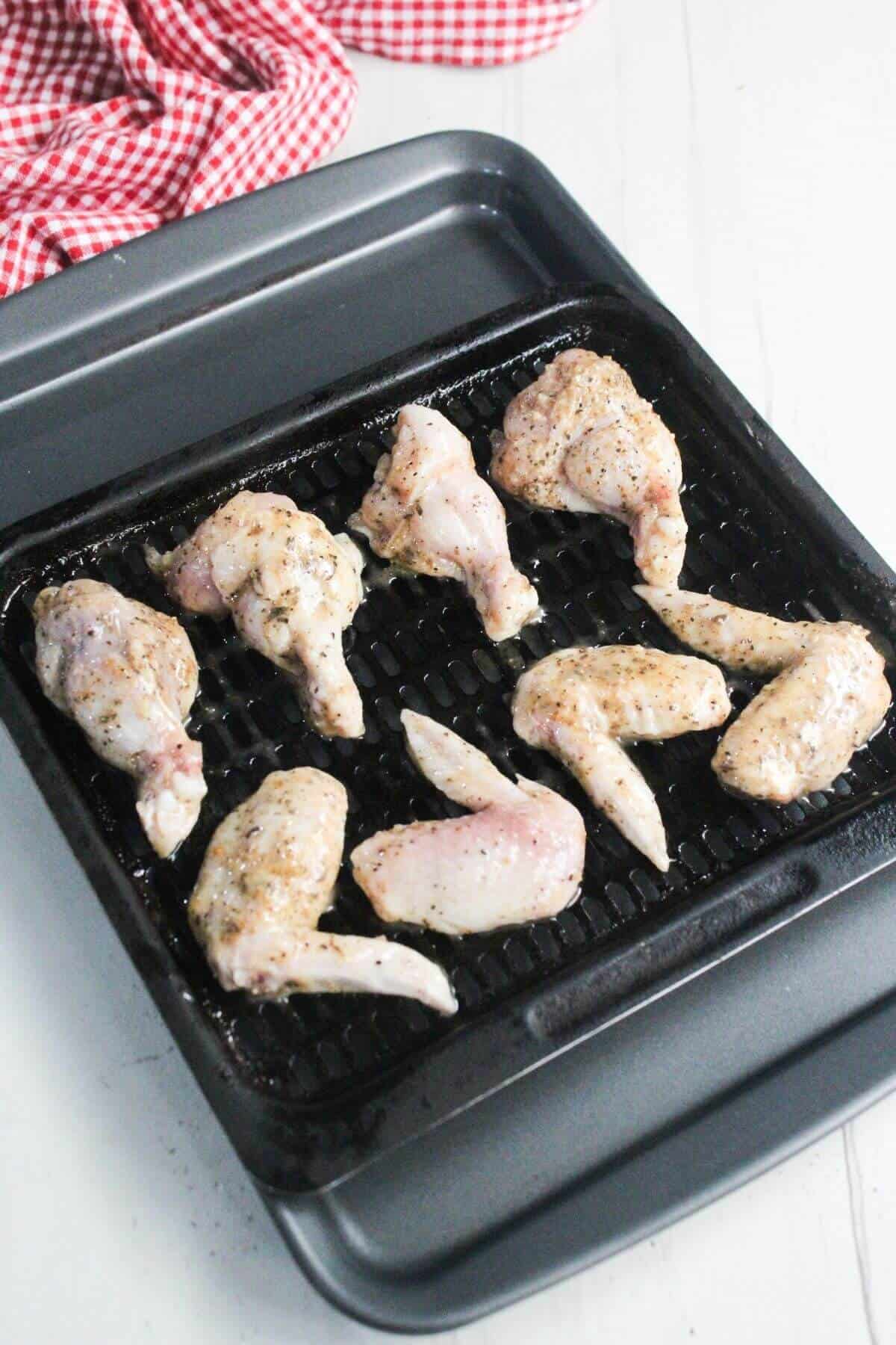 Raw seasoned chicken wings arranged on a black grill pan, ready to be cooked. A red and white checkered cloth is in the background.