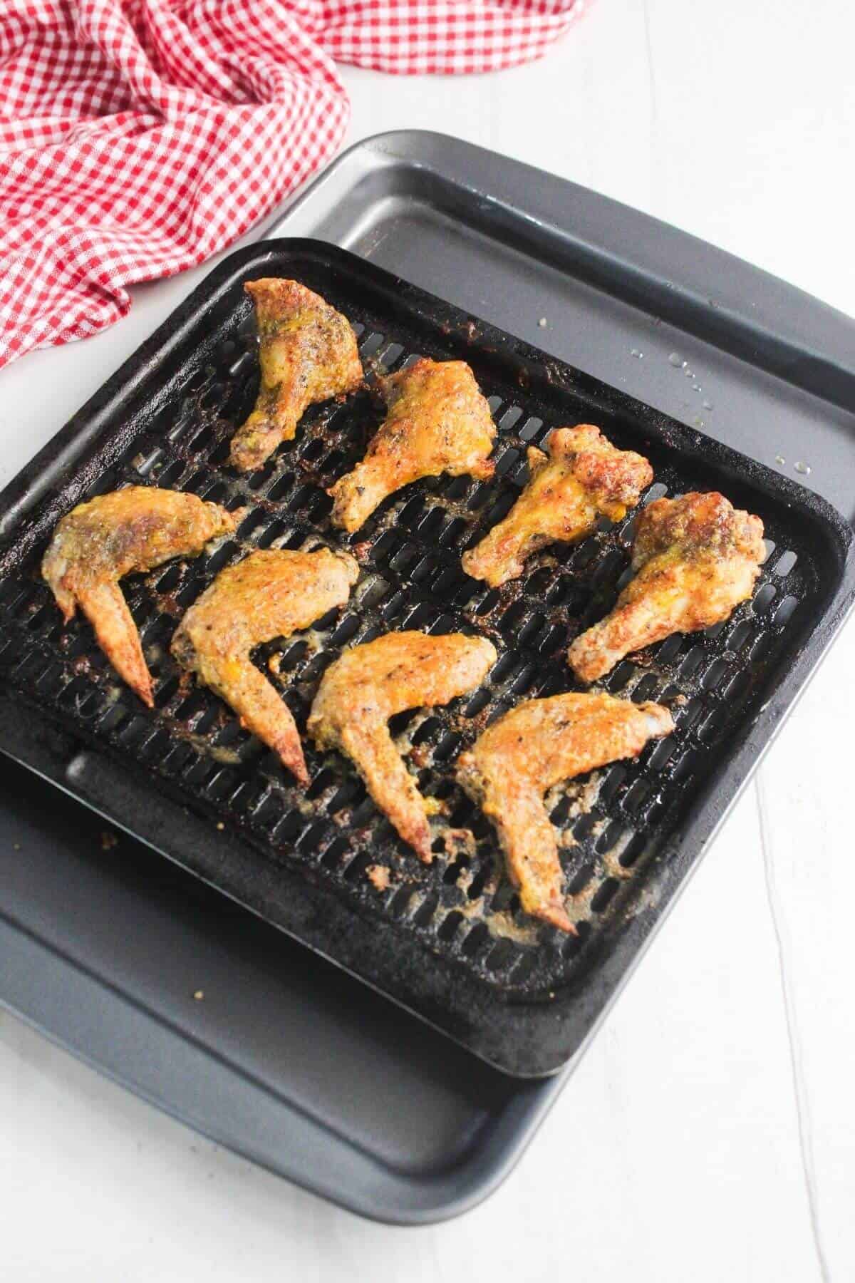 Eight cooked chicken wings on a grill pan with a baking tray underneath, placed on a white surface with a red and white checkered cloth nearby.